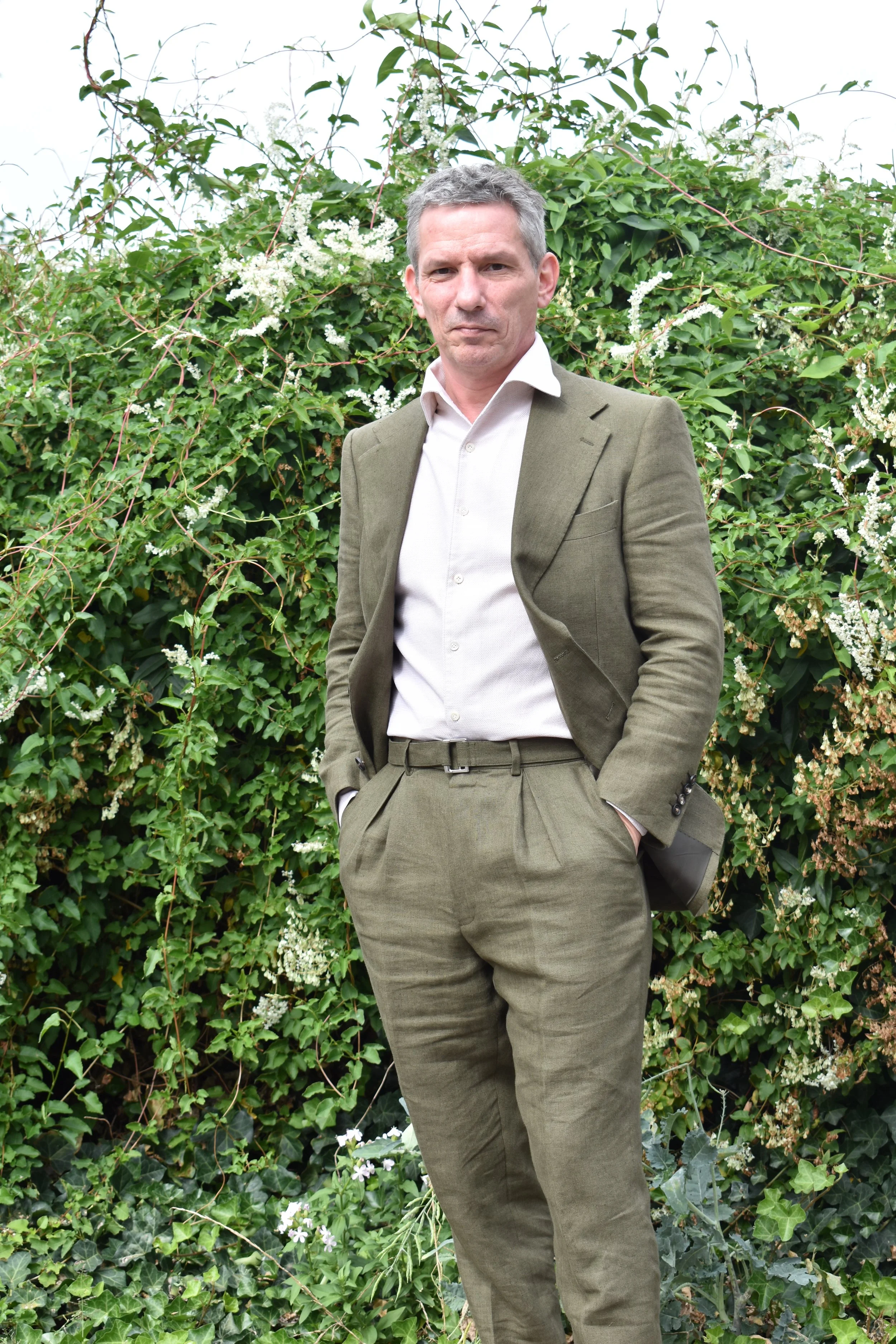 A man wearing a green suit and white shirt standing outdoors in front of a bush with white flowers and green leaves.