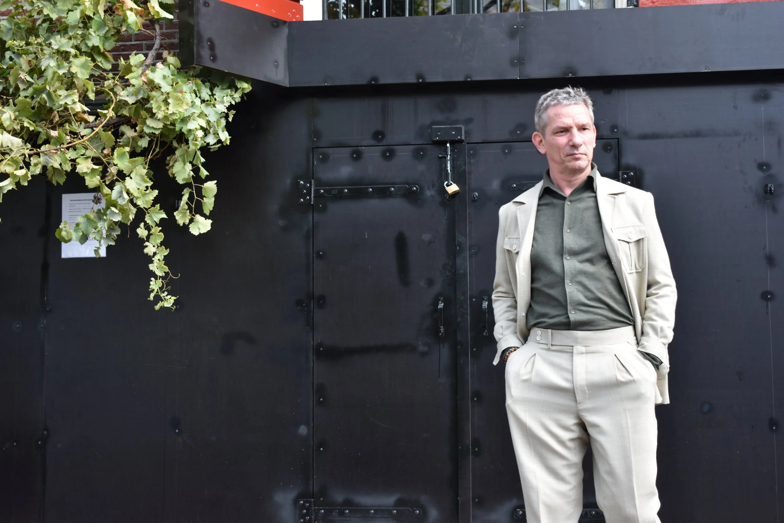 A middle-aged man in a beige suit jacket and pants with a green shirt stands with hands in his pockets against a dark wall, with green leaves hanging on the left side of the image.