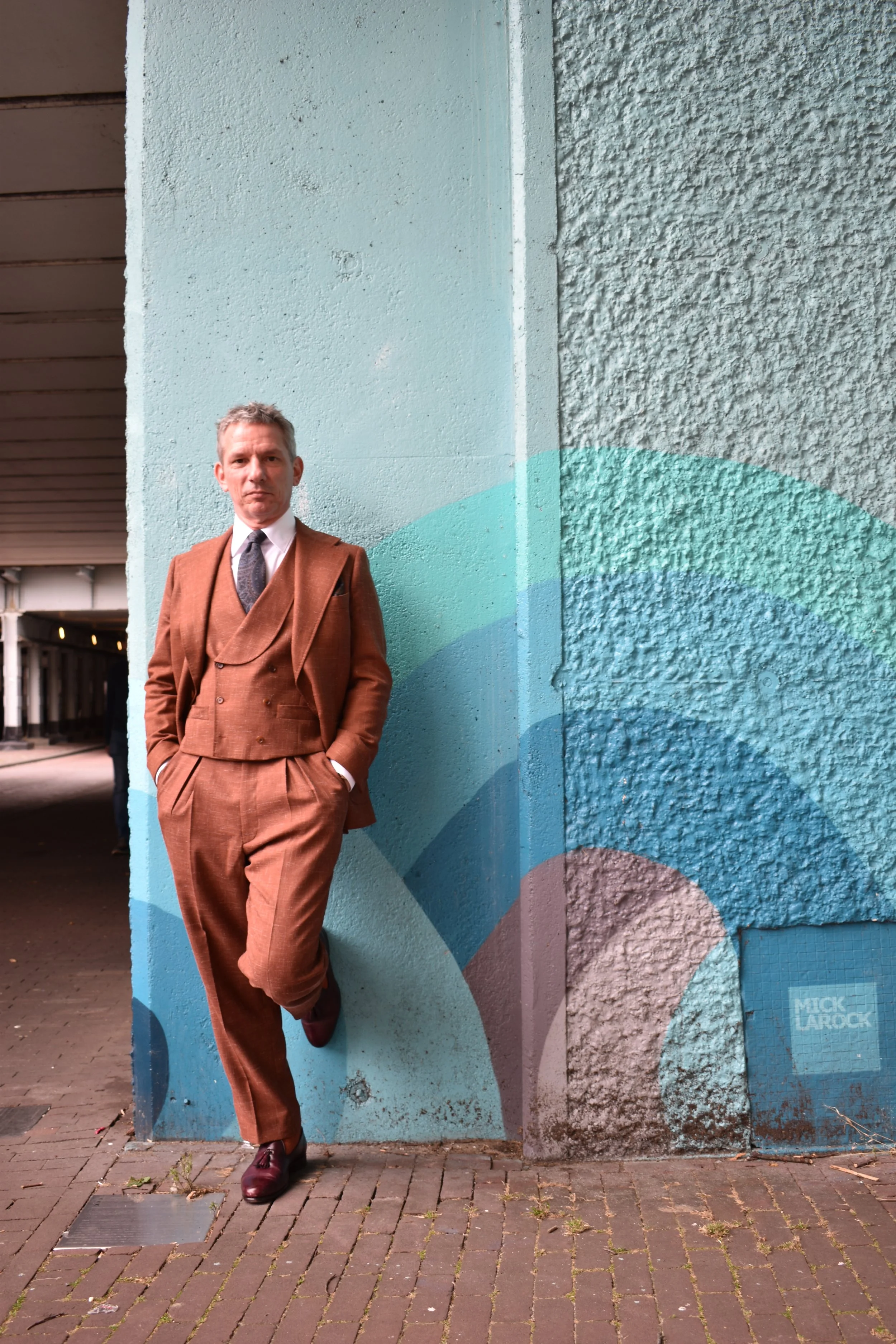 A man in a brown suit with a double-breasted vest and tie leaning against a colorful abstract mural on a wall.