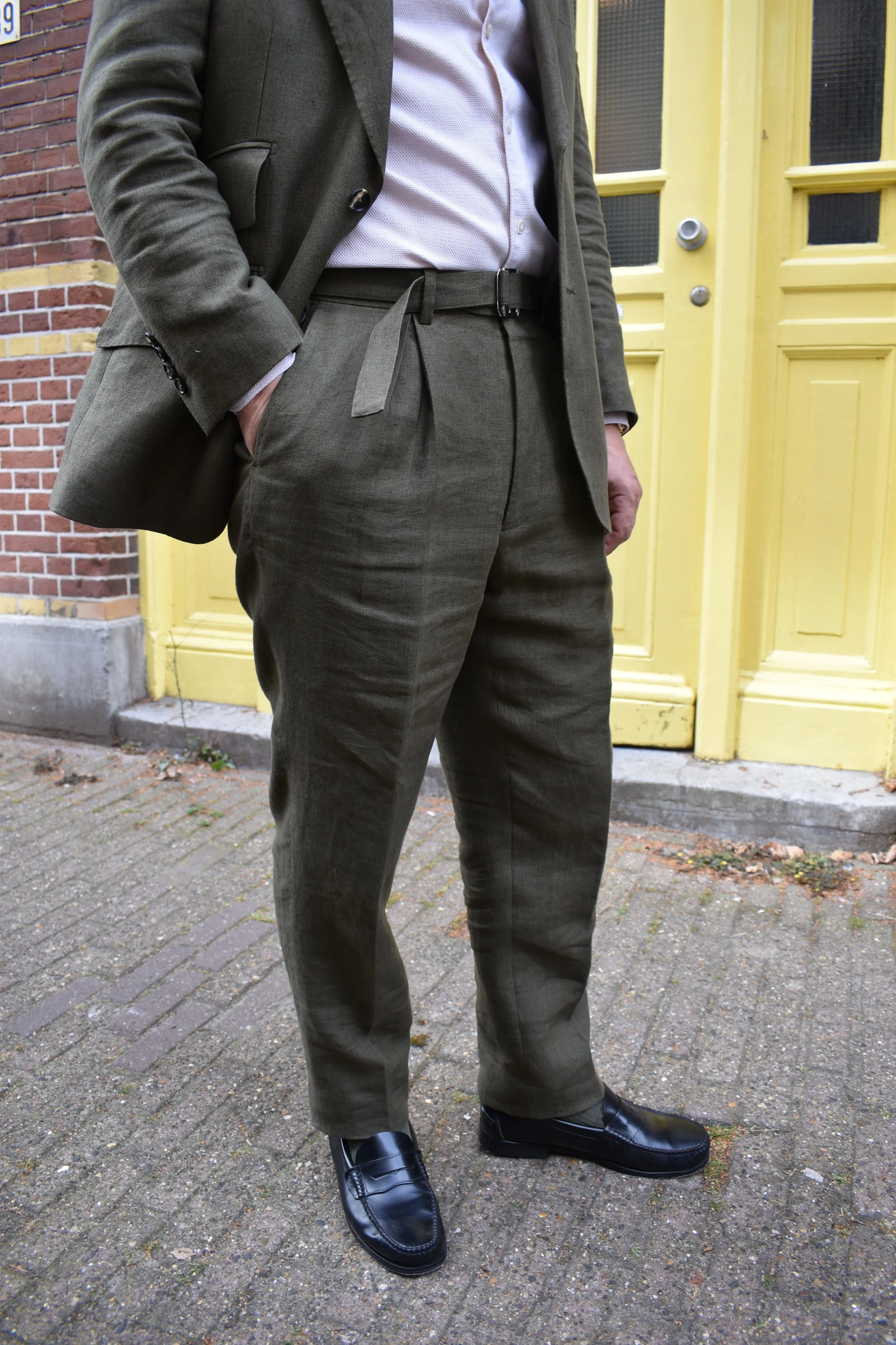 Person dressed in a dark gray suit with a light-colored button-up shirt, standing outside near a yellow door.