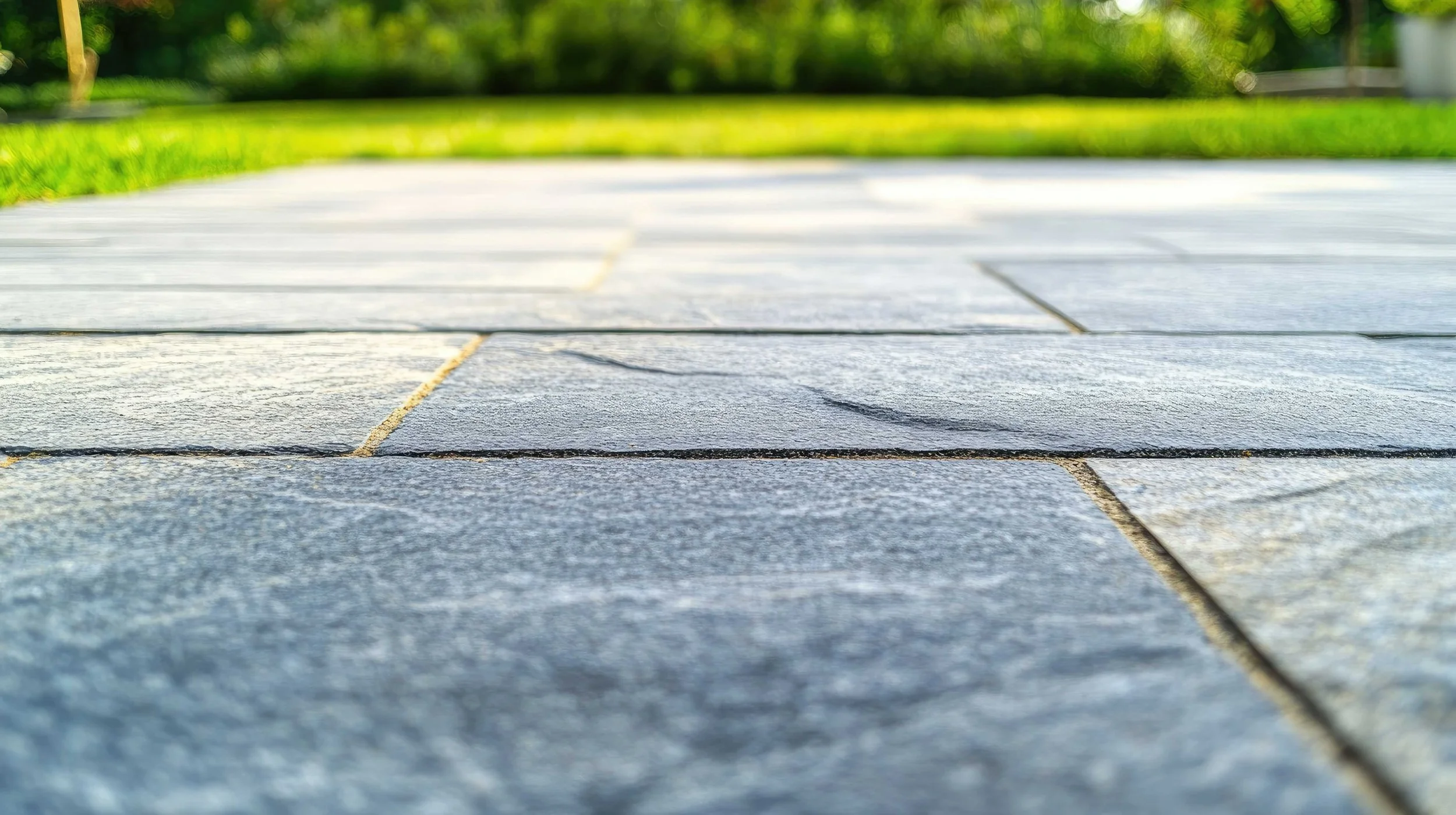 Close-up view of gray stone pavement with cracks, with a background of green grass and trees under bright sunlight.
