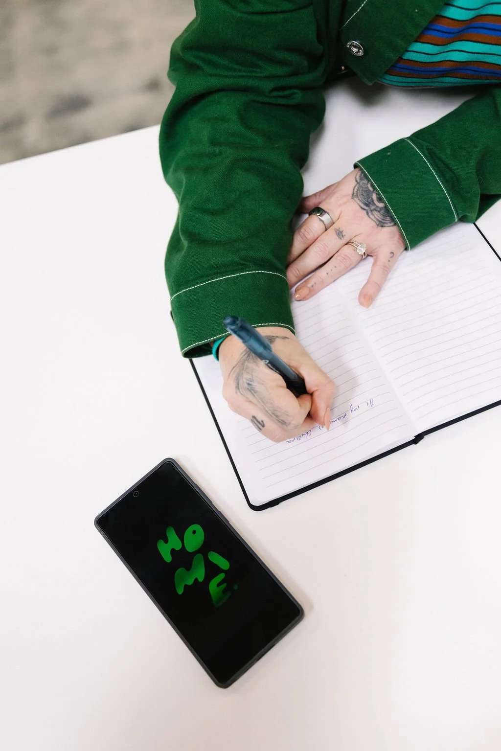 A person with tattoos on their hand is writing in a lined notebook with a black pen. They are wearing a green shirt or jacket and have multiple rings on their fingers. There is a smartphone on the white table, displaying green letters on its screen.