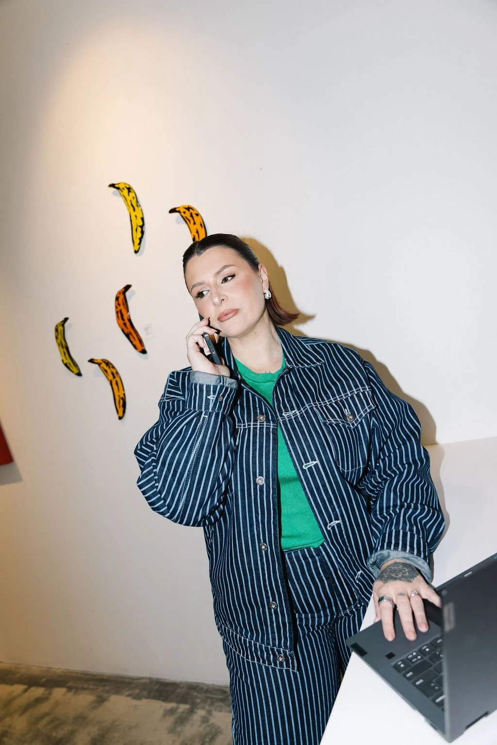 A woman with short brown hair wearing a pinstripe jacket and green shirt, talking on a phone, in an art gallery with banana-shaped decorations on the wall.