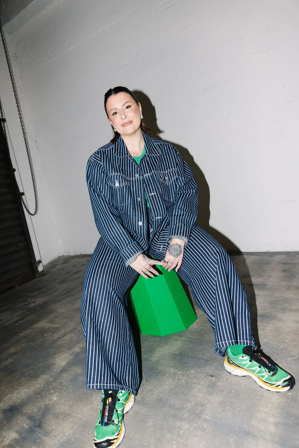 A woman with dark hair, wearing a pinstripe matching jacket and pants, sitting on a green cube in a room with white walls and concrete floor. She wears green sneakers and has tattoos on her left hand.