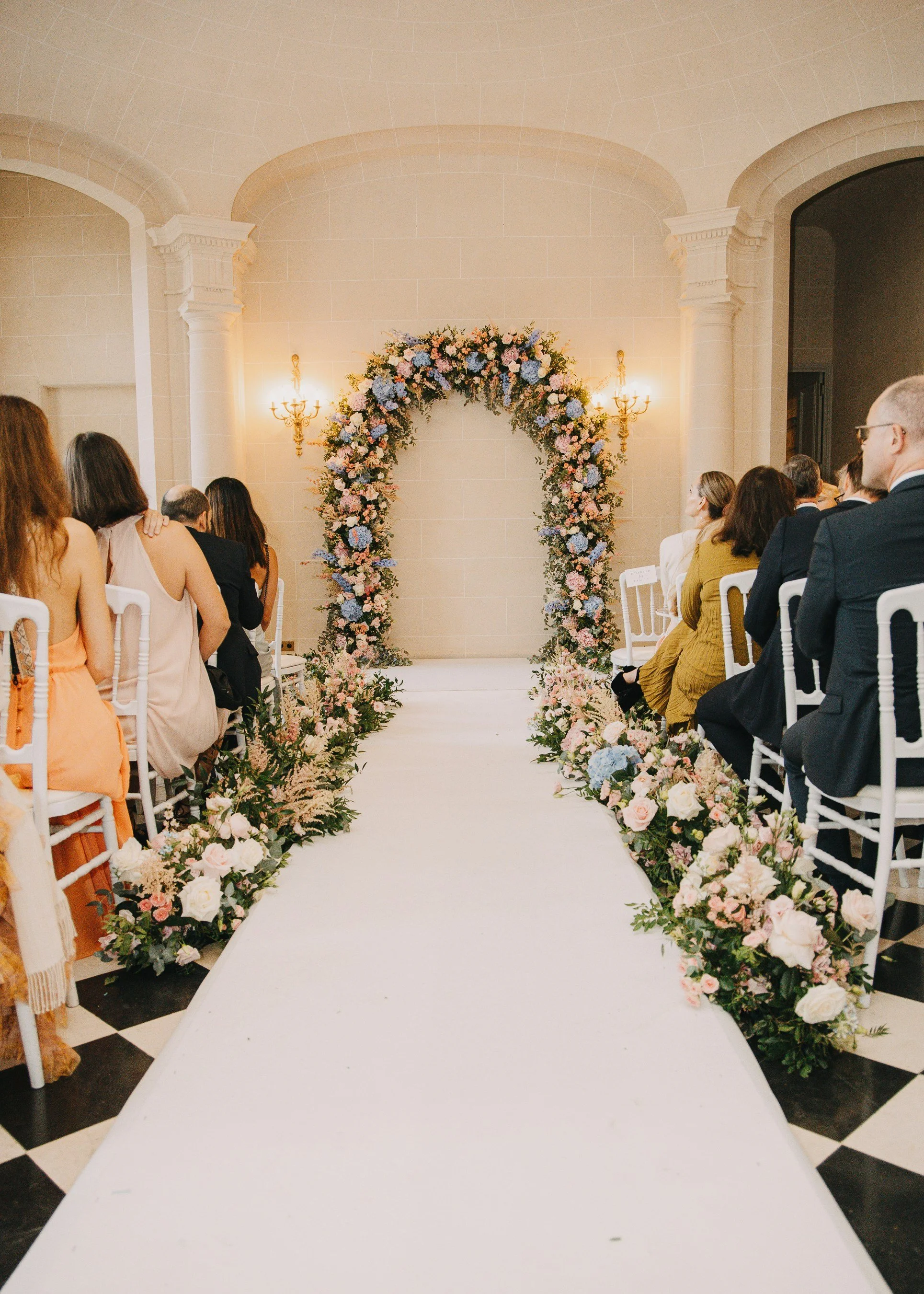 Hochzeitsdekoration mit Blumenbogen und Blumendekoration entlang des Gangs in einem festlichen Raum mit weißen Stühlen und Anwesenden auf beiden Seiten.