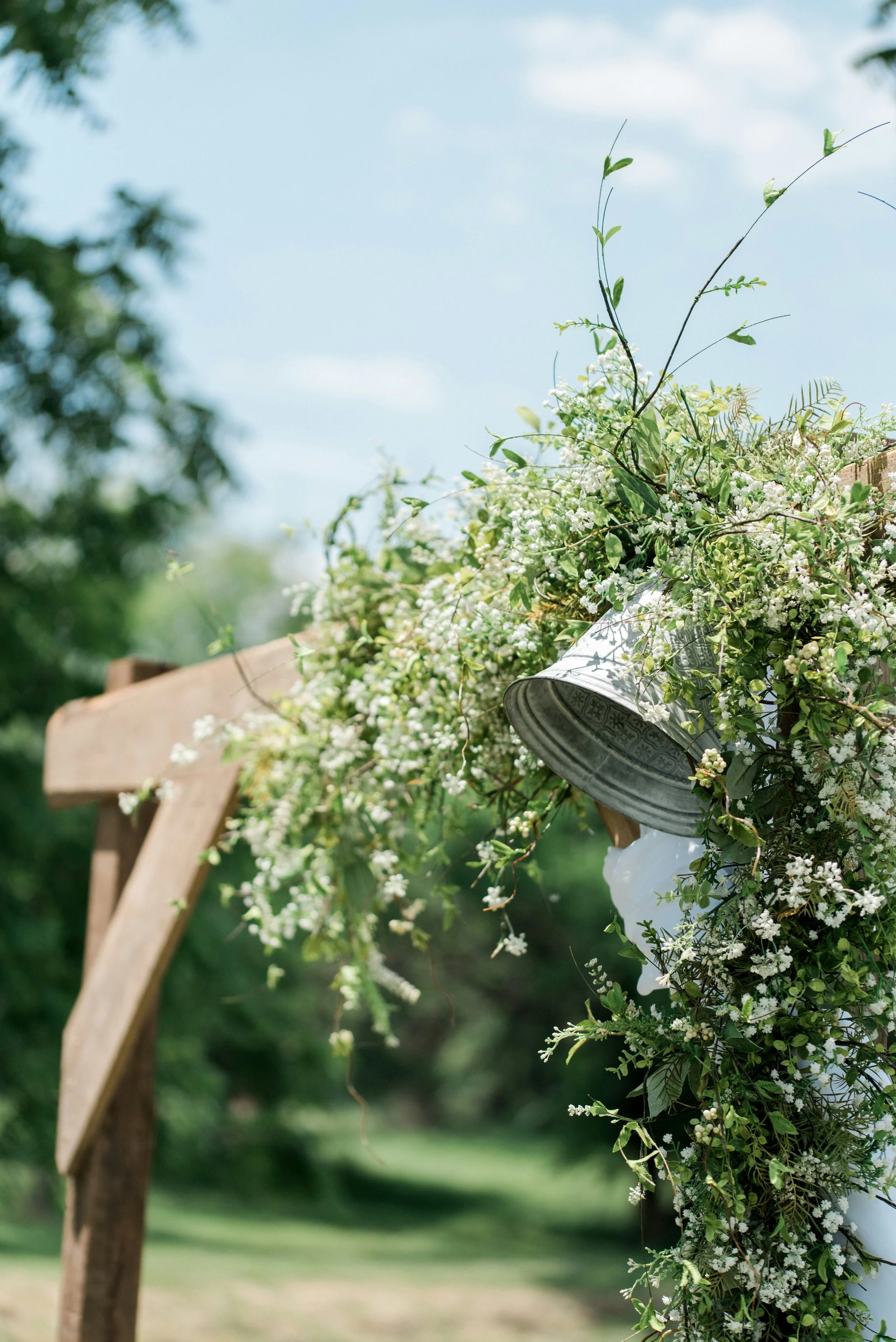 Blumen- und Pflanzenarrangements an einem Holzkreuz bei einer Hochzeit im Freien, mit blauer Himmel im Hintergrund.