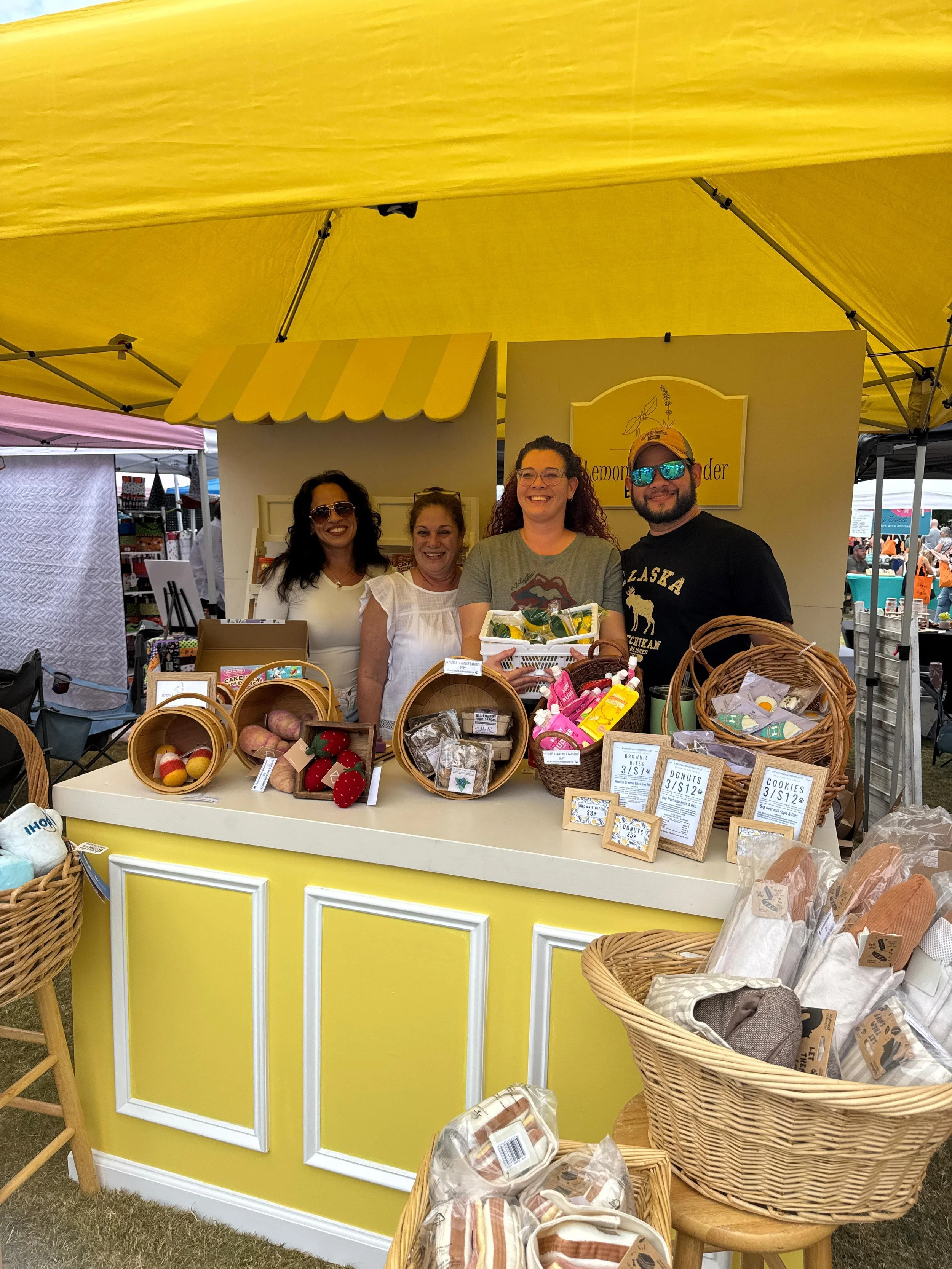 The Lemons & Lavender Barkery team smiling behind their yellow market booth, surrounded by artisan dog treats and curated gift baskets at a local festival in the Atlanta area
