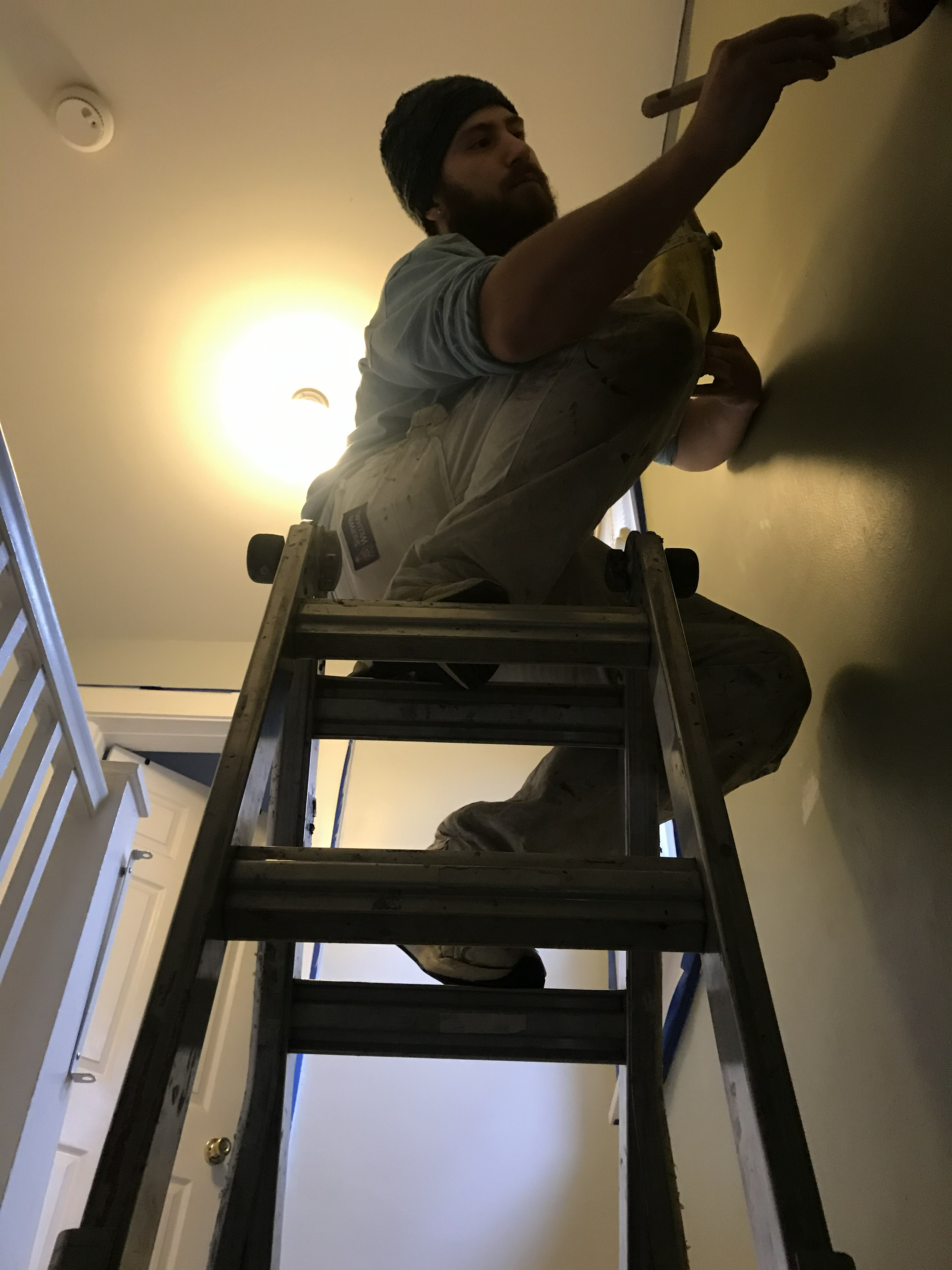 A man in work clothes, sitting on a ladder, painting a wall in a room with a ceiling light on.