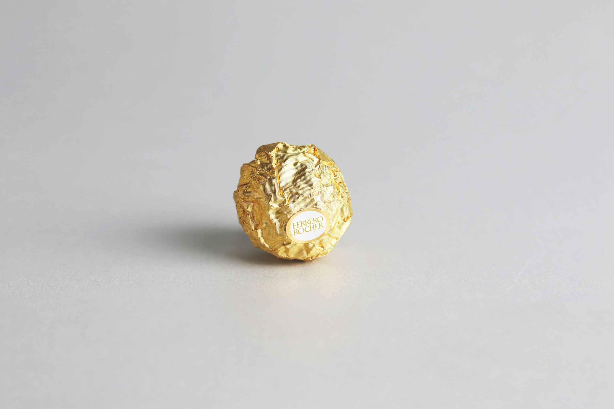 A single Ferrero Rocher chocolate wrapped in gold foil on a gray background.