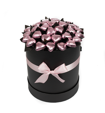 A black round box decorated with a pink ribbon bow, filled with heart-shaped chocolates wrapped in pink foil.