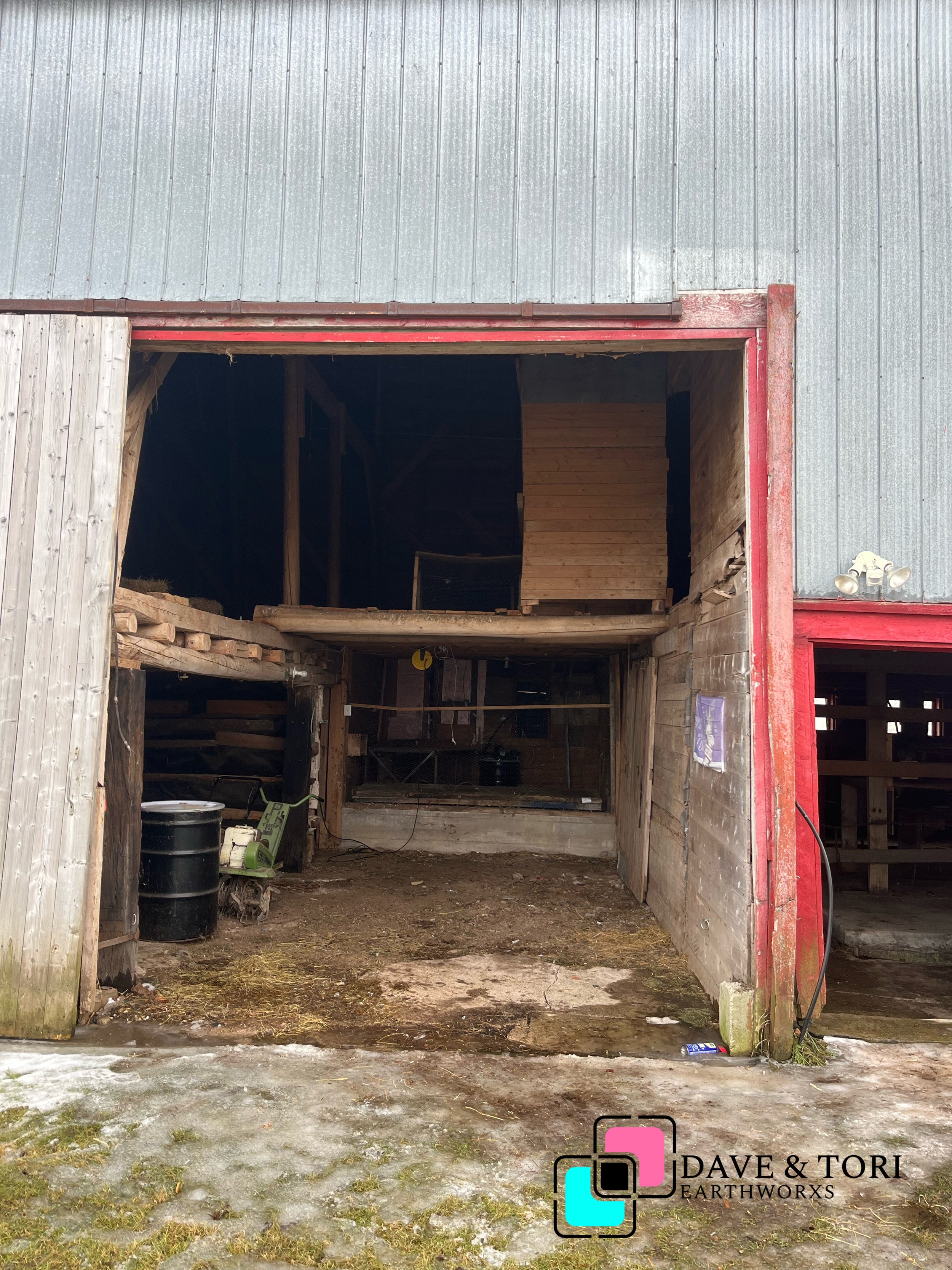 The image shows the front of a partially constructed or renovated barn or shed with open large doorway. The ground outside is dirt with patches of snow and moss. Inside the barn, there are construction tools, debris, and wooden structures. The exterior walls are made of corrugated metal and wood, with some areas showing red framing. A logo for DAVE & TORI EARTHWORXS is in the bottom right corner.