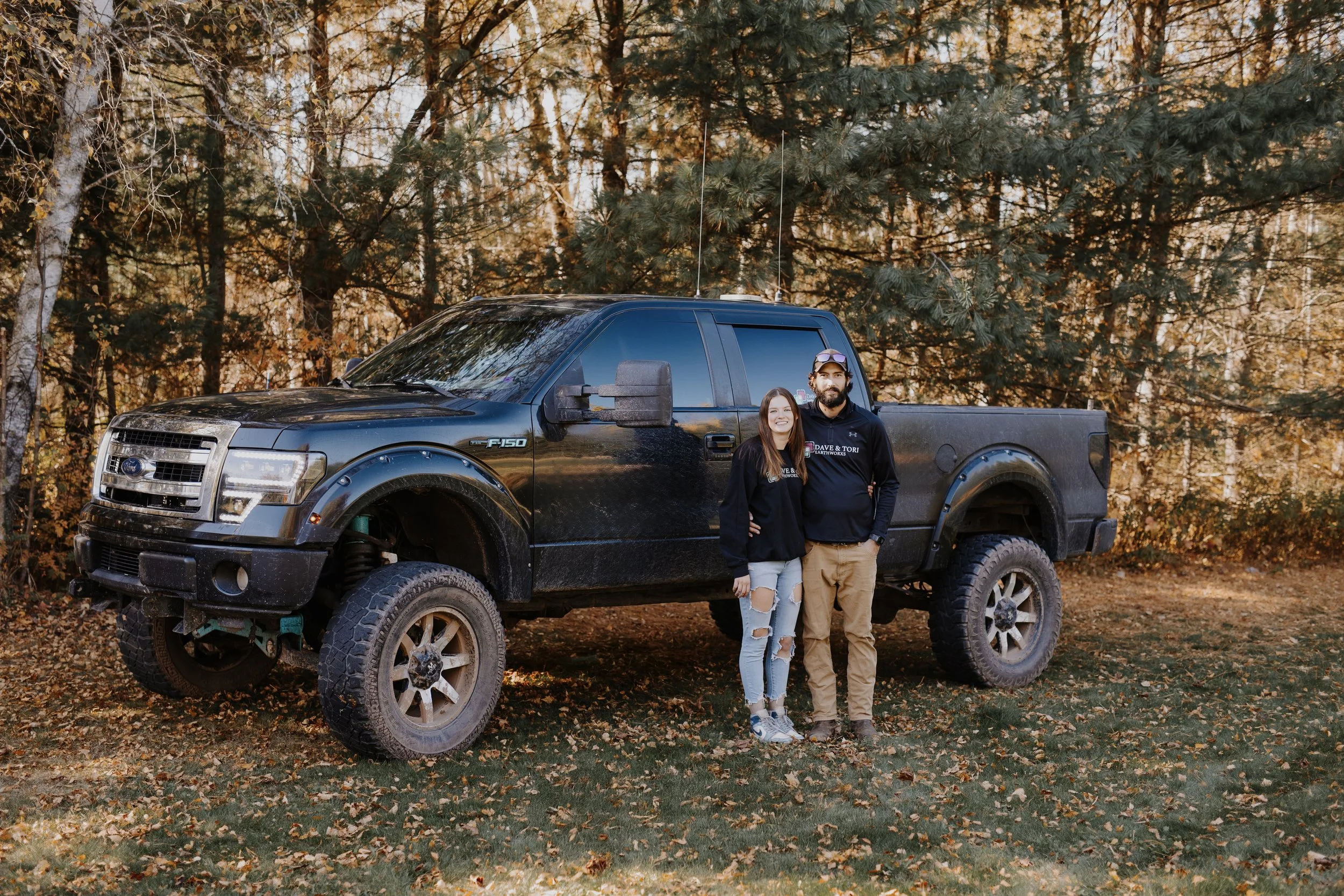 A couple standing in front of a black Ford F-150 truck in a wooded area with autumn leaves.