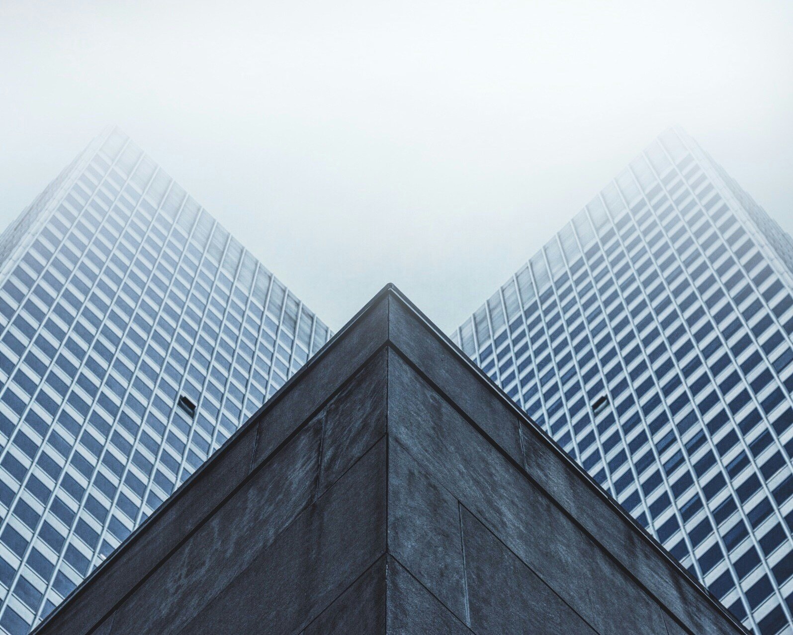 Low-angle view of modern glass office buildings with a contrasting dark stone building corner in the foreground, against an overcast sky.