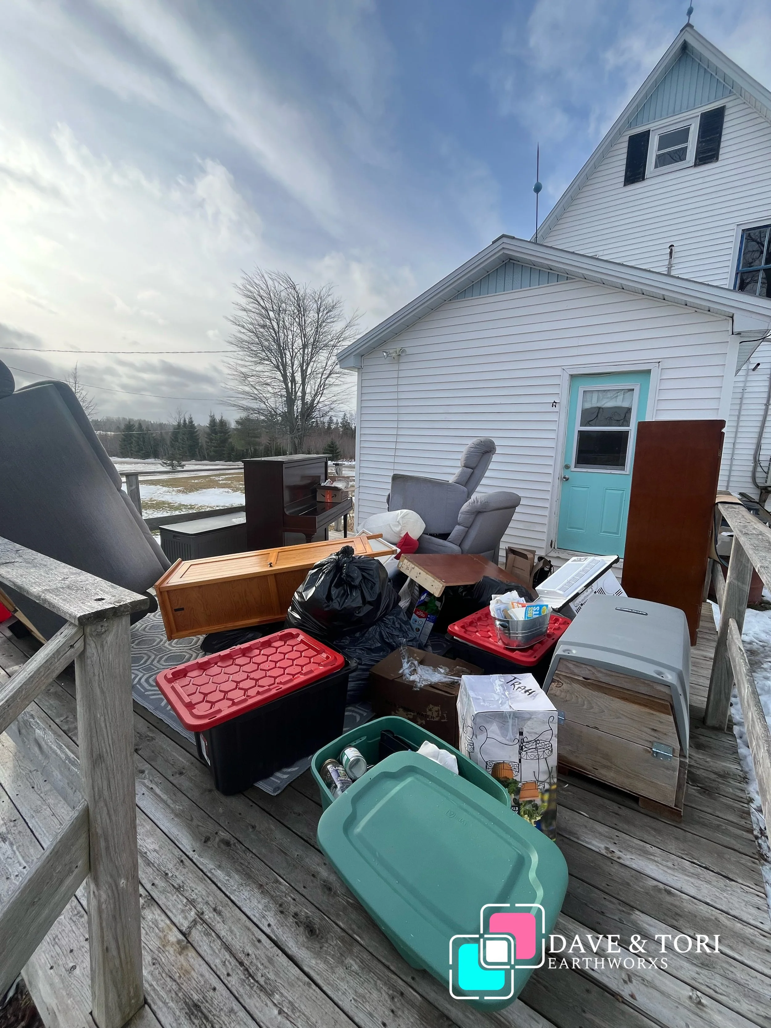 House with a cluttered wooden deck filled with various items including boxes, bins, chairs, and furniture, set against a partly cloudy sky.