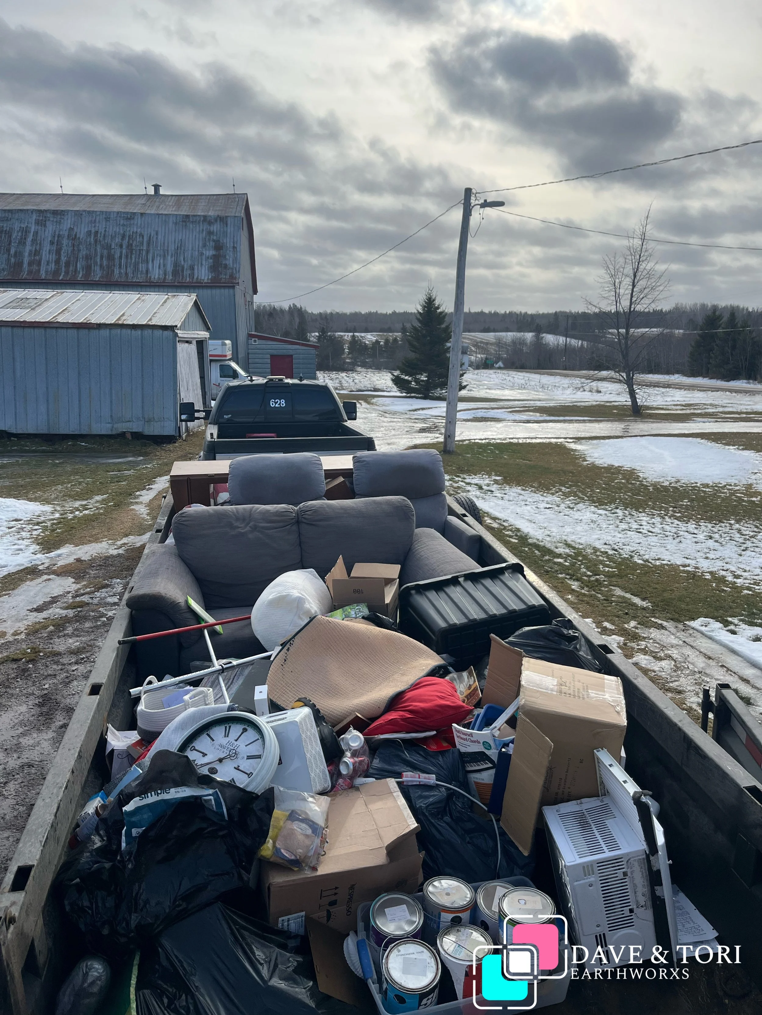 A trailer filled with various household items, including a clock, cardboard boxes, a black toolbox, a black plastic bag, a white lantern, and other miscellaneous objects, parked outdoors on a cold day with patches of snow on the ground.