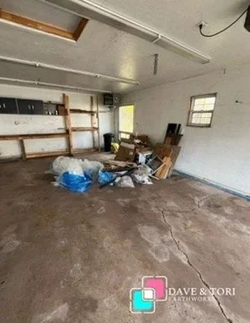 Empty garage with concrete floor, some scattered trash and cardboard, and wooden shelves against the wall.