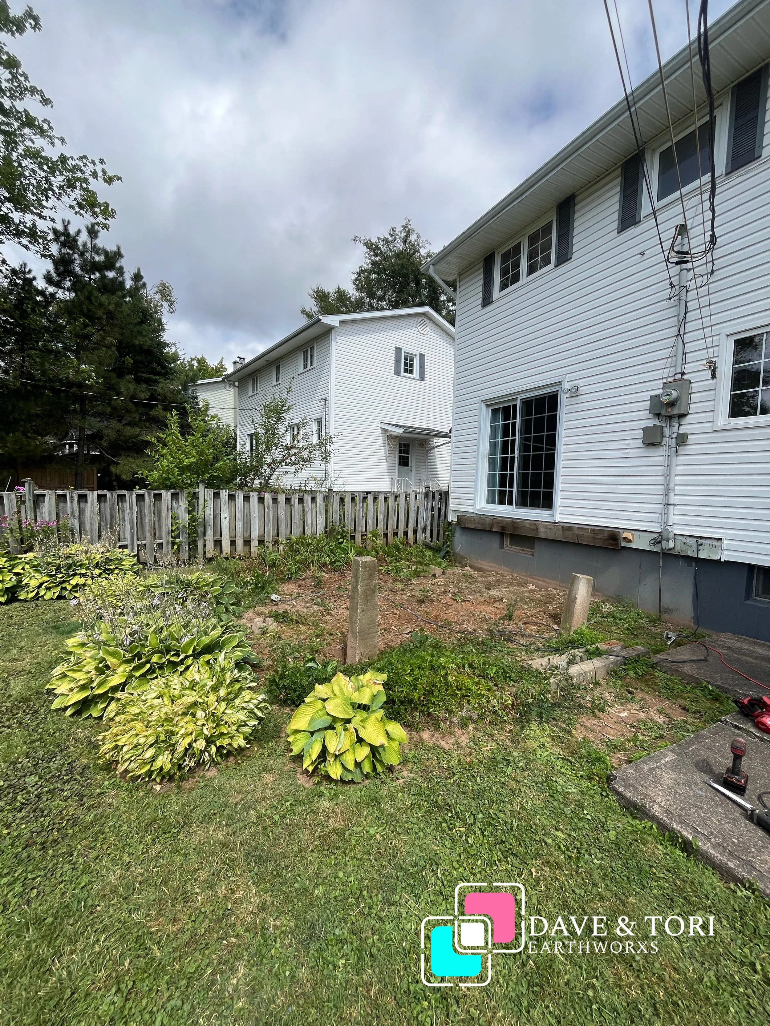 Backyard of a house with landscaping, including hostas and a small garden bed, and a wooden fence. There are two houses with white siding visible in the background under a cloudy sky. The yard has some construction or repair work in progress, with concrete blocks and tools present, along with an electrical meter on the house. The logo for 'Dave & Tori Earthworxs' is visible in the bottom right corner.