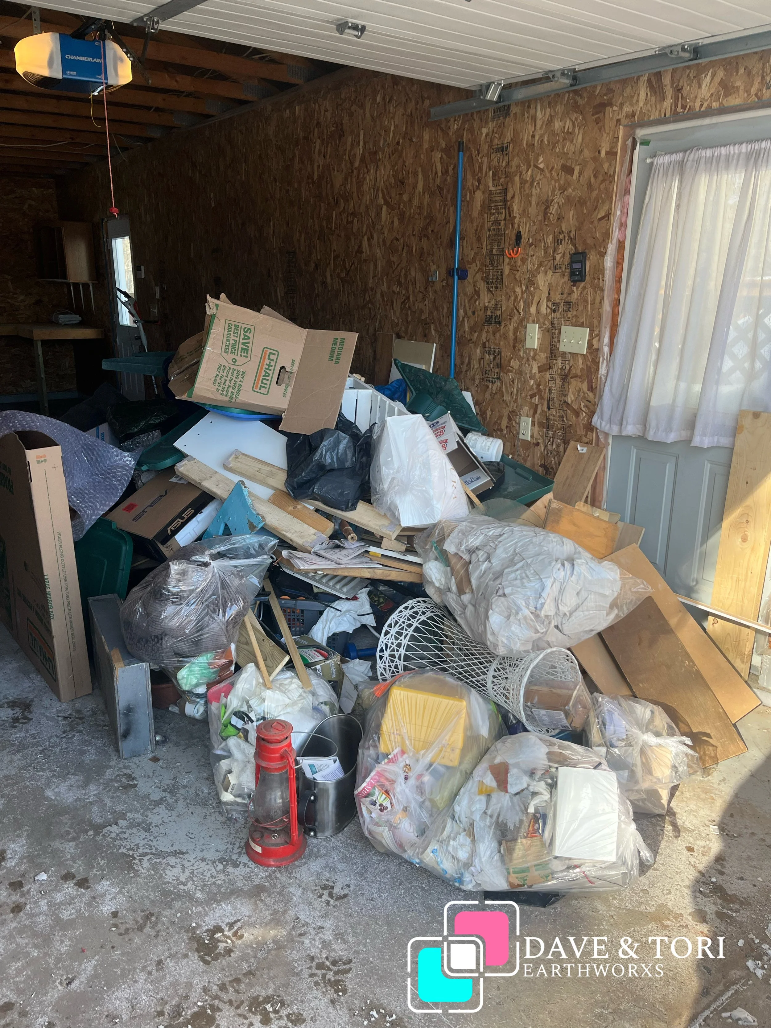 A cluttered garage with piles of trash, cardboard boxes, and miscellaneous items on the floor. There is a garage door on the right side, window with curtains, and wood-paneled walls.