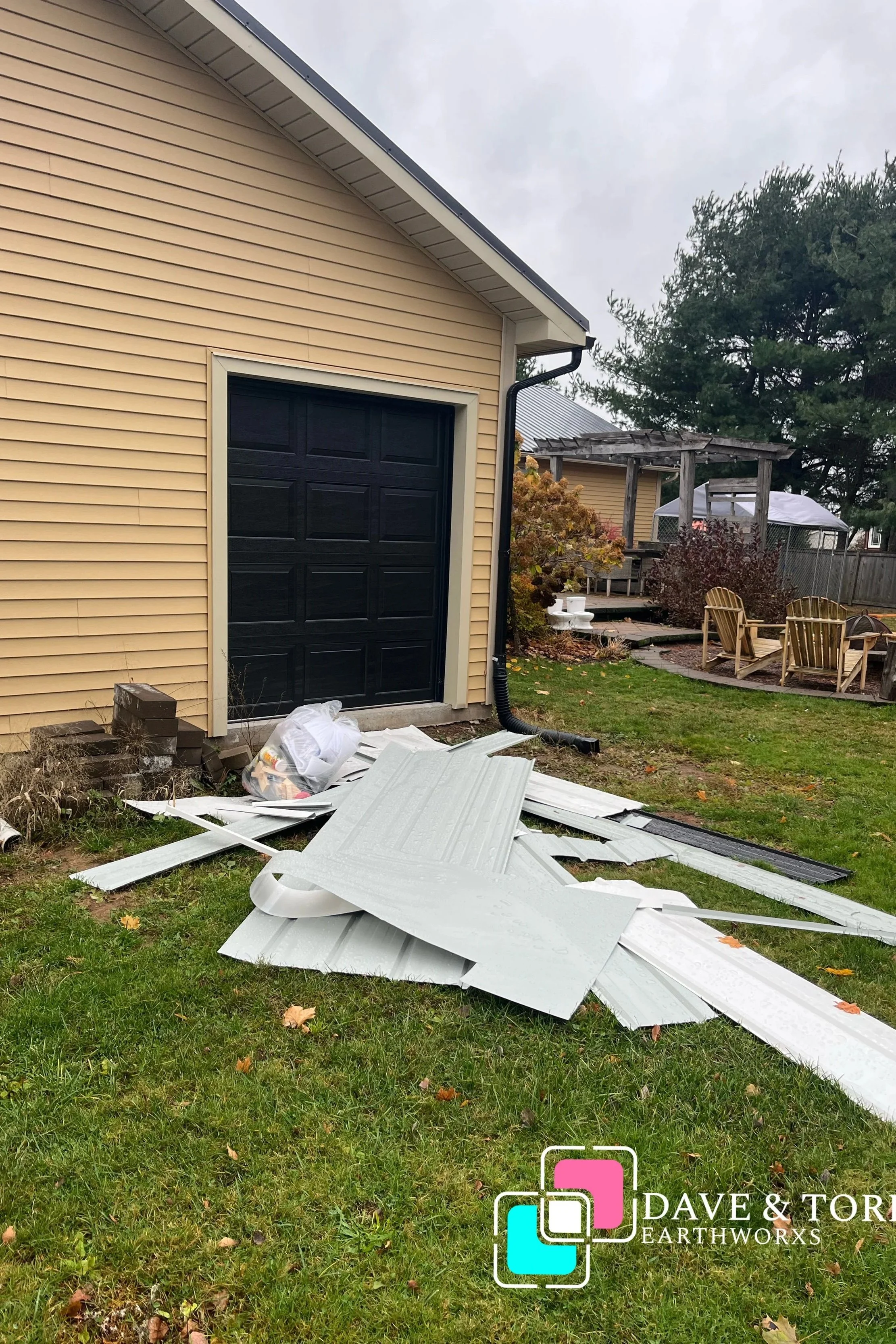 Debris and fallen siding panels on a lawn in front of a beige garage with a black door, rain droplets visible on the panels, and a plastic bag filled with trash nearby. In the background, a backyard with outdoor seating, trees, and a small deck can be seen.