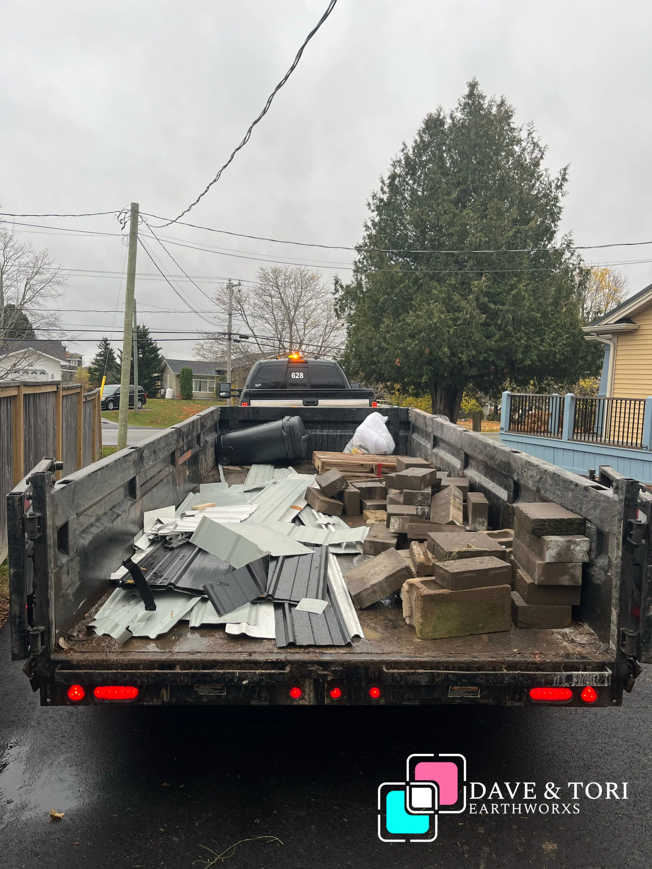 Back of a flatbed truck loaded with construction debris including metal roofing sheets, bricks, and a black trash can. Residential neighborhood with trees and houses in the background, overcast sky, wet pavement.