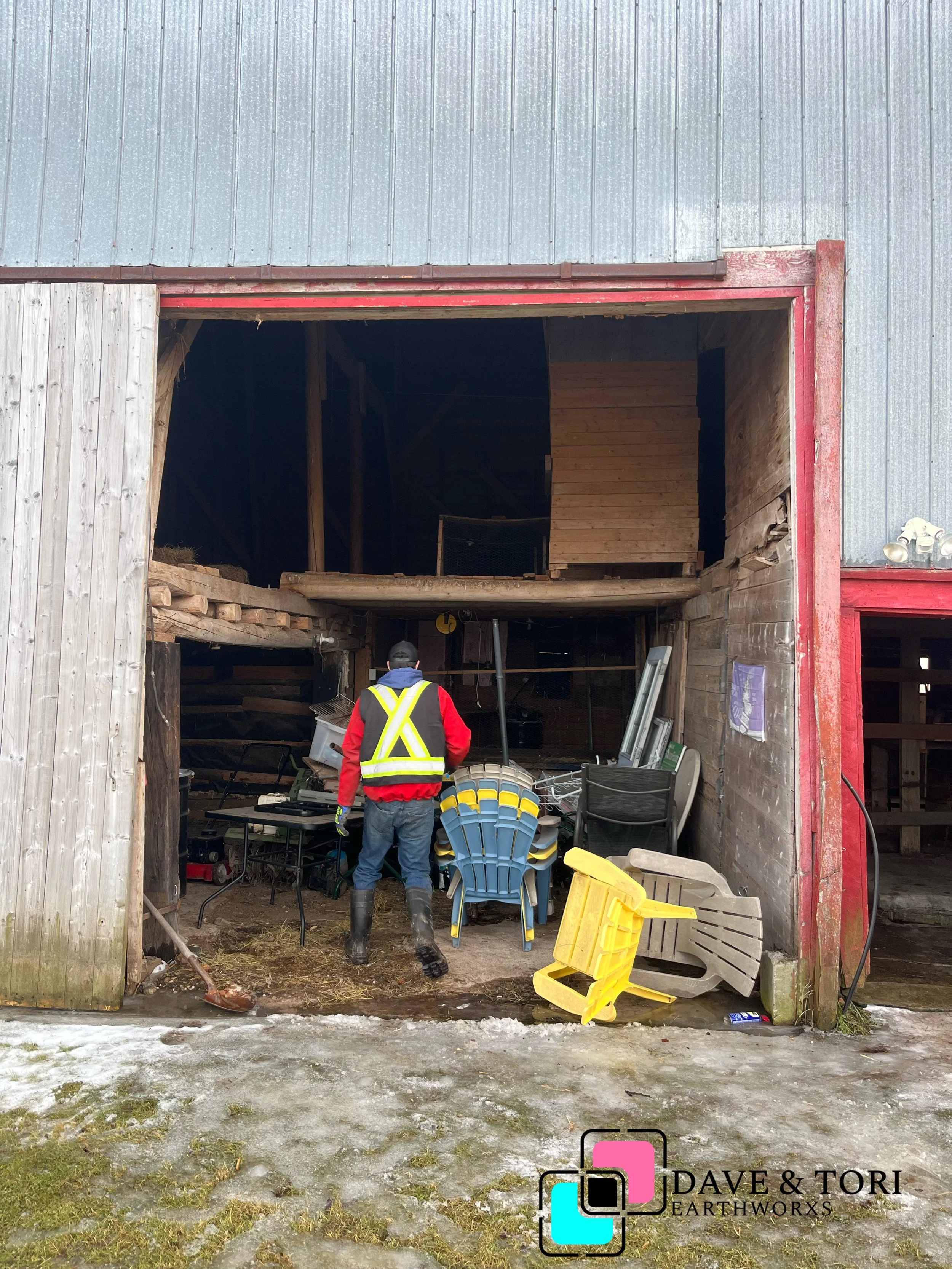 A person wearing a red jacket, black hat, and black boots stands inside an open barn, surrounded by stacked plastic chairs and miscellaneous items, with a partially visible icy ground outside.