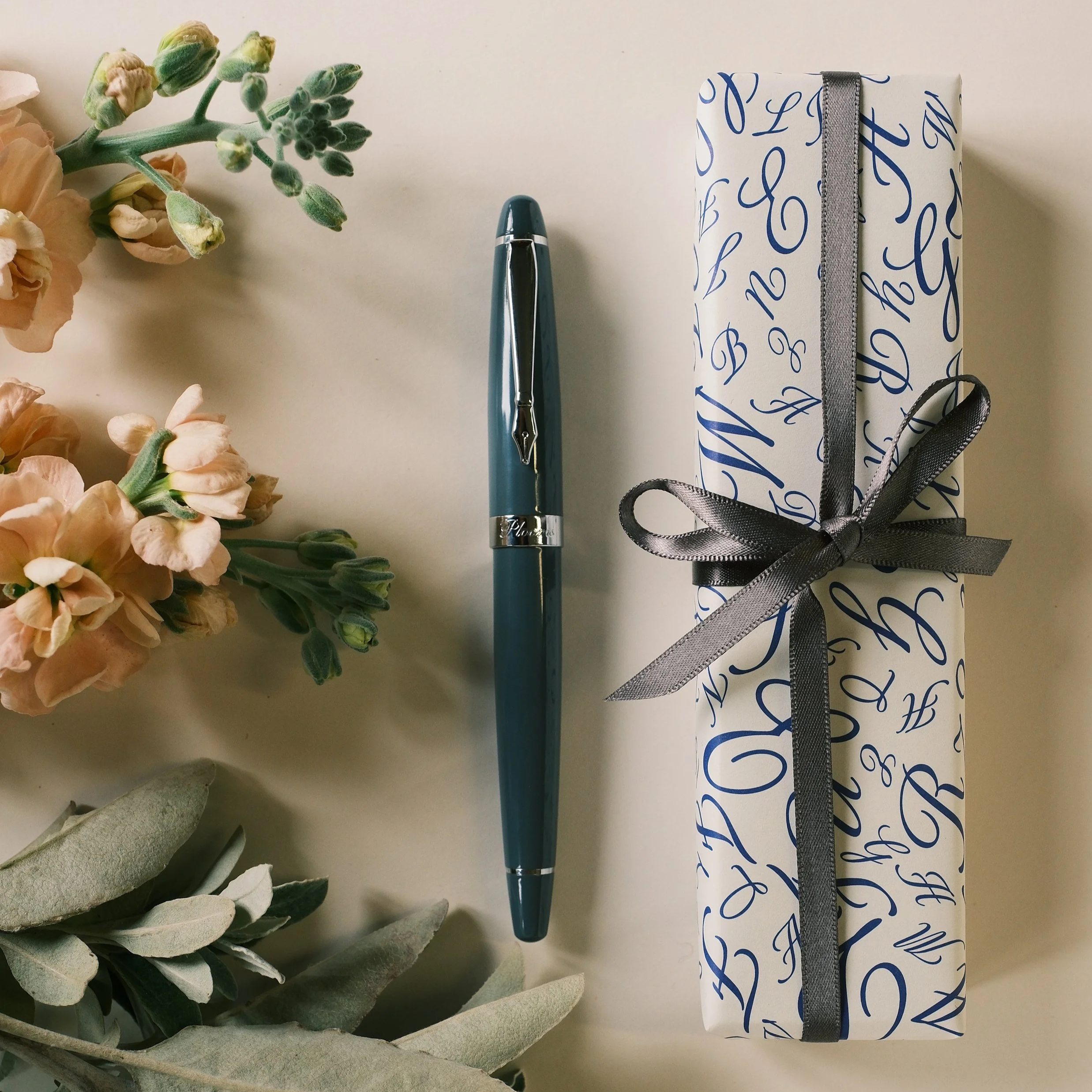 A blue pen with silver accents, a wrapped gift box with blue cursive lettering tied with a black ribbon, and pink peony flowers and green foliage arranged on a light surface.