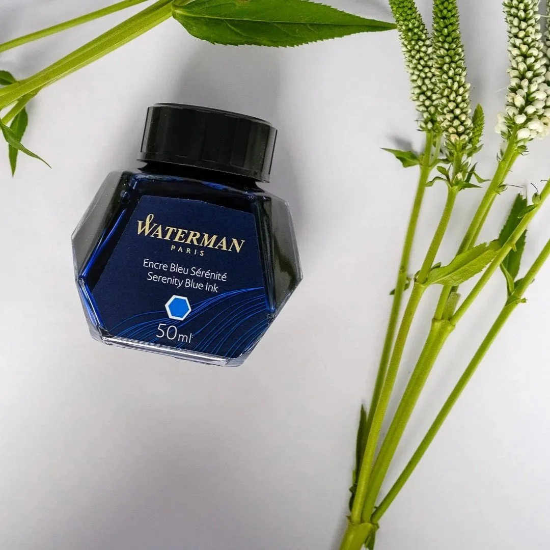 A blue ink bottle labeled 'Waterman Paris Serenity Blue Ink, 50ml' next to some green leaves and white flower spikes on a white background.