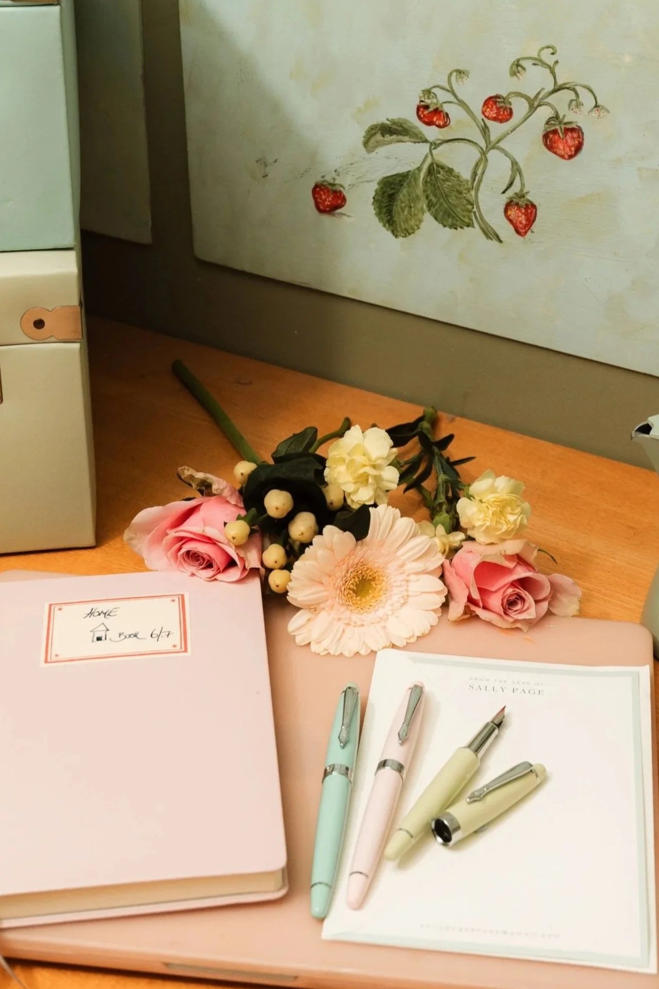 A desk with a pink notebook, three pastel-colored pens, a notepad, a small bouquet of pastel flowers, and a framed picture of strawberries on the wall.