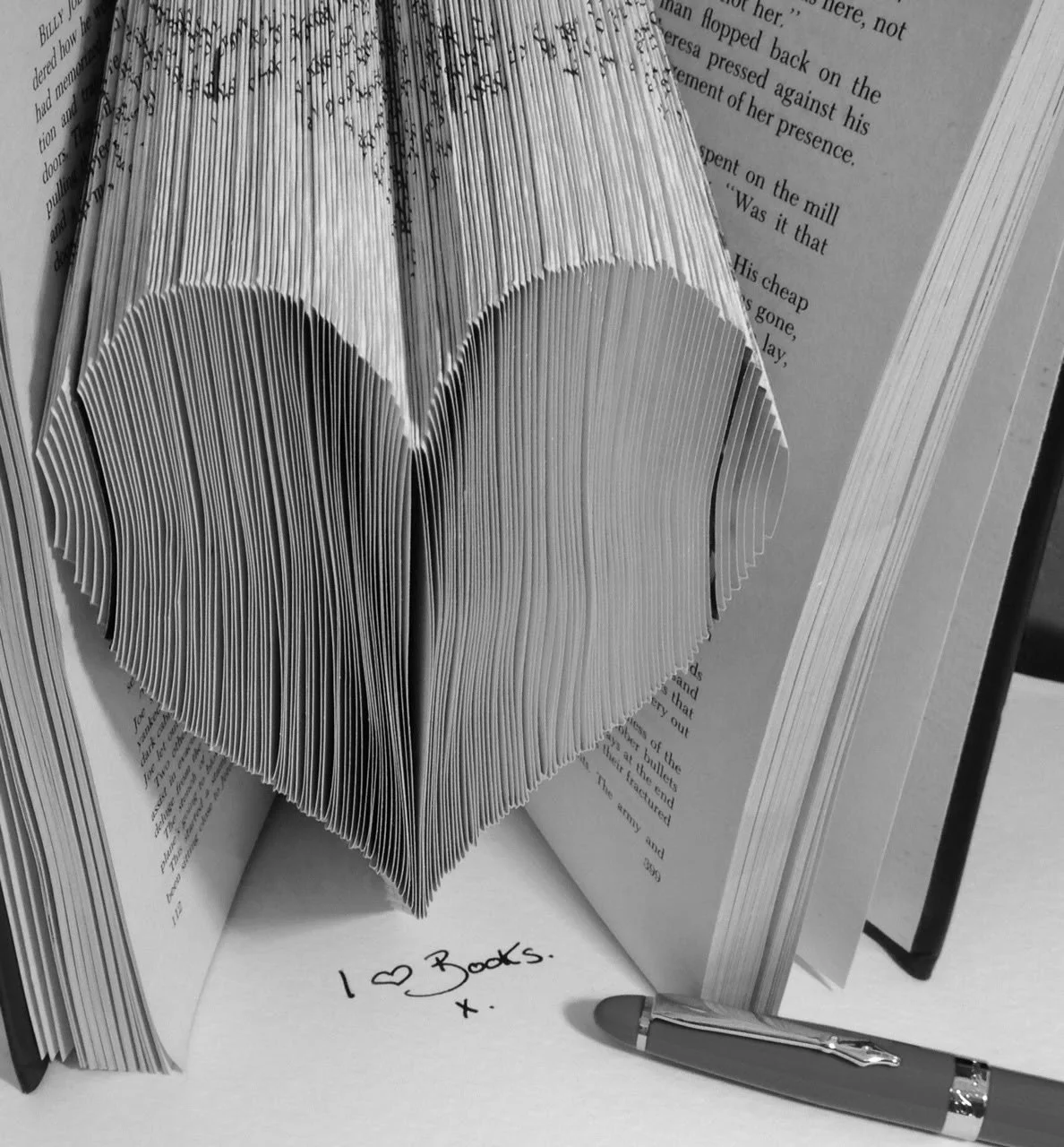 An open book with pages folded to create a heart shape. A handwritten note on a piece of paper in front of the book reads "I love books. x" and a pen is also visible.