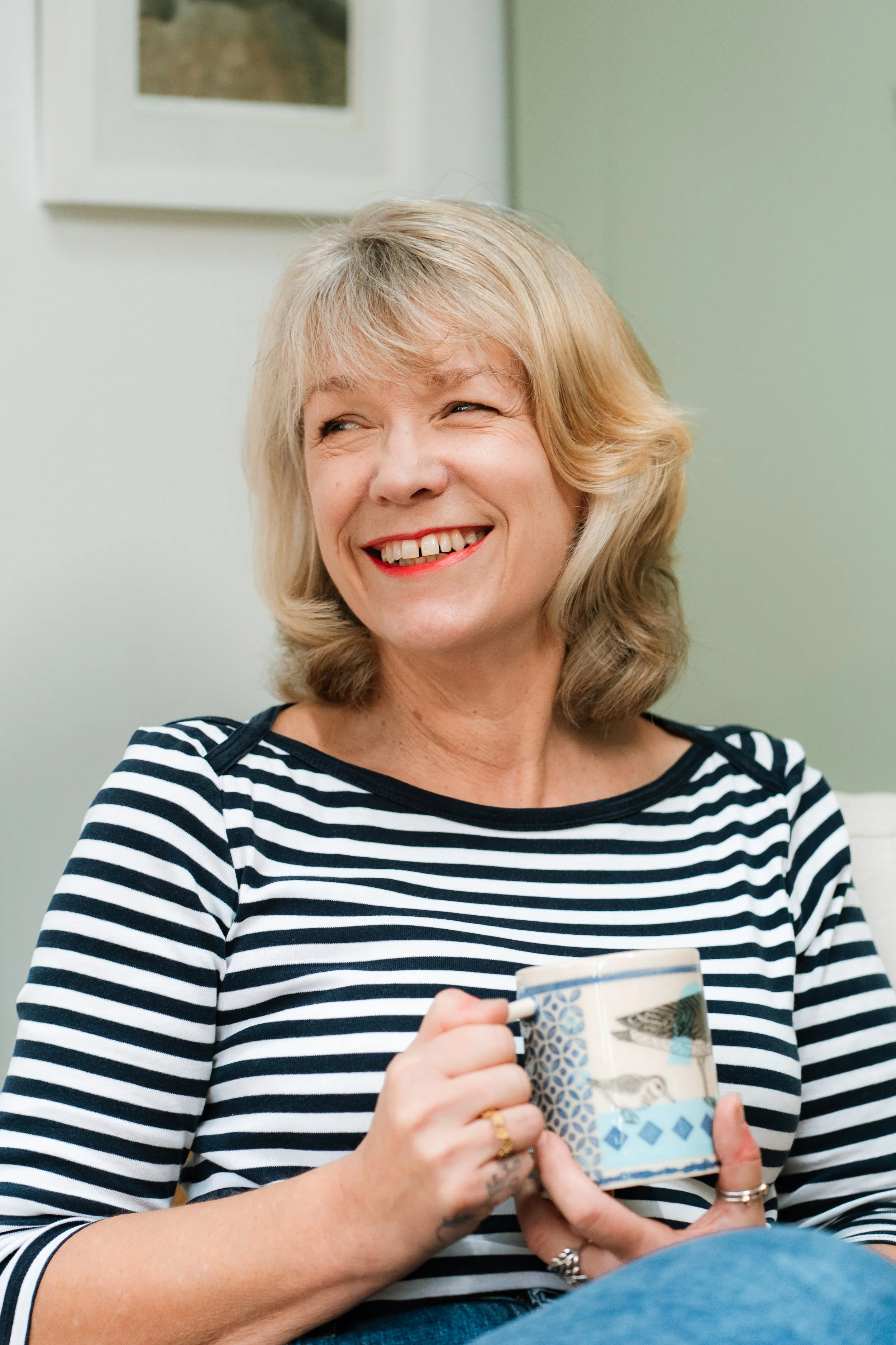 A smiling woman with blonde hair holding a colorful mug, wearing a black and white striped shirt, sitting in a cozy room with artwork on the wall.