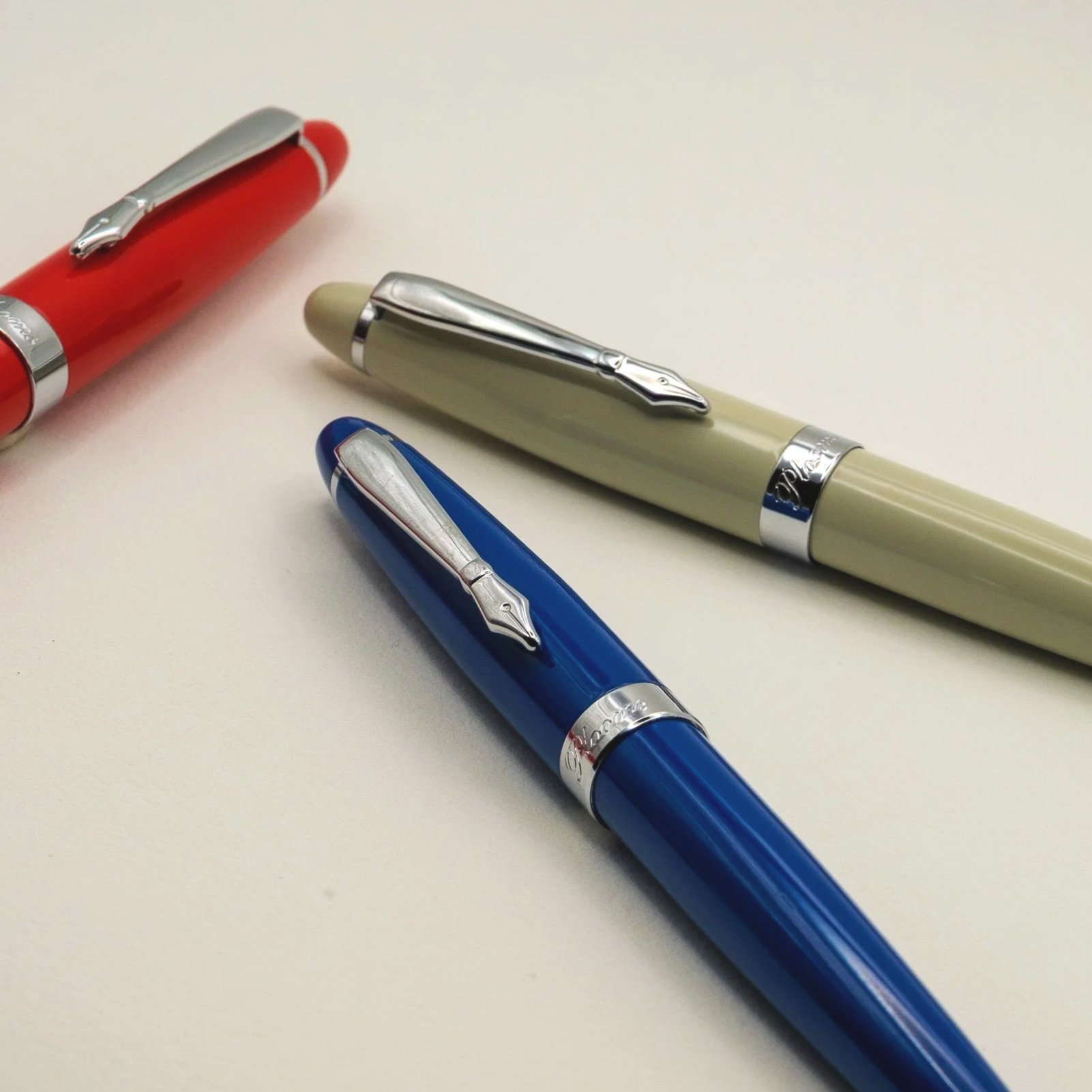 Three fountain pens in red, cream, and blue on a white surface.