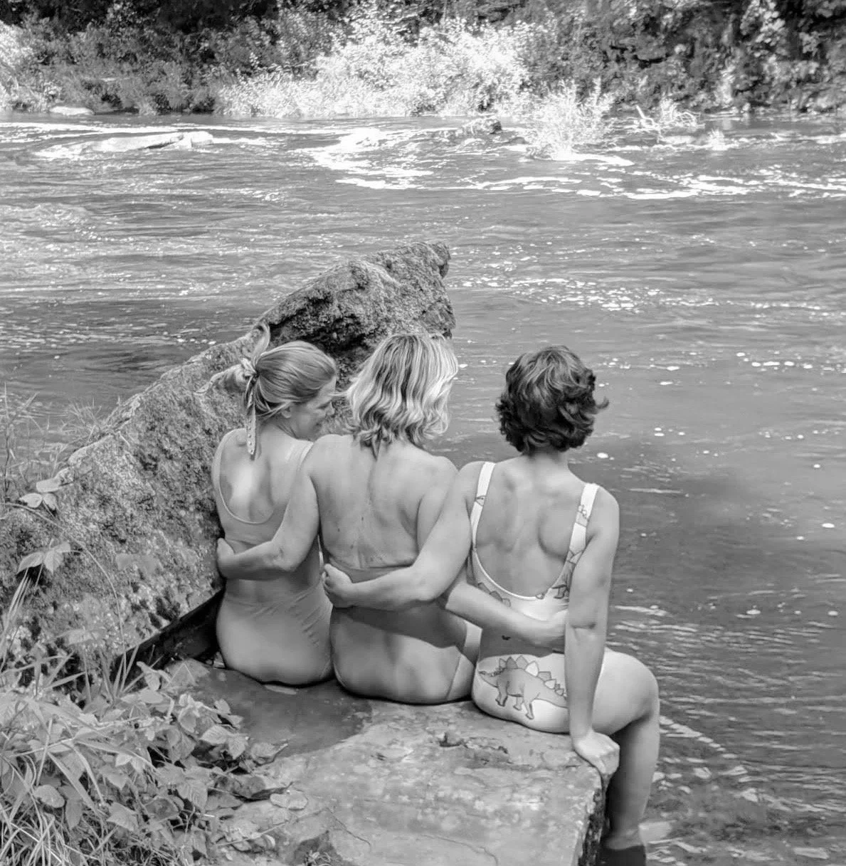 Three women sitting on a rock by the water, wearing swimsuits, with their backs to the camera, looking at the water and waves in the distance.