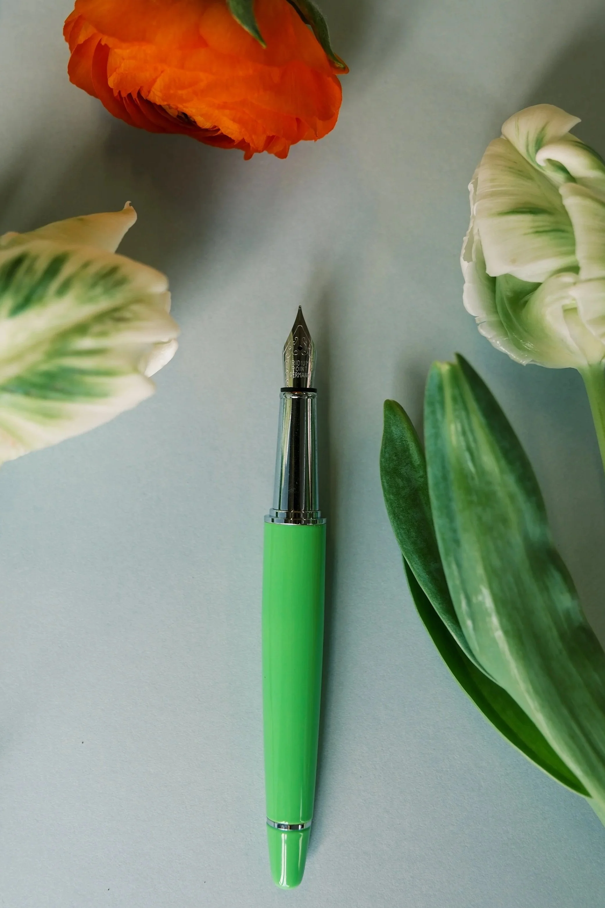 A green fountain pen with a silver nib, placed on a light-colored surface surrounded by orange, white, and green tulip flowers.