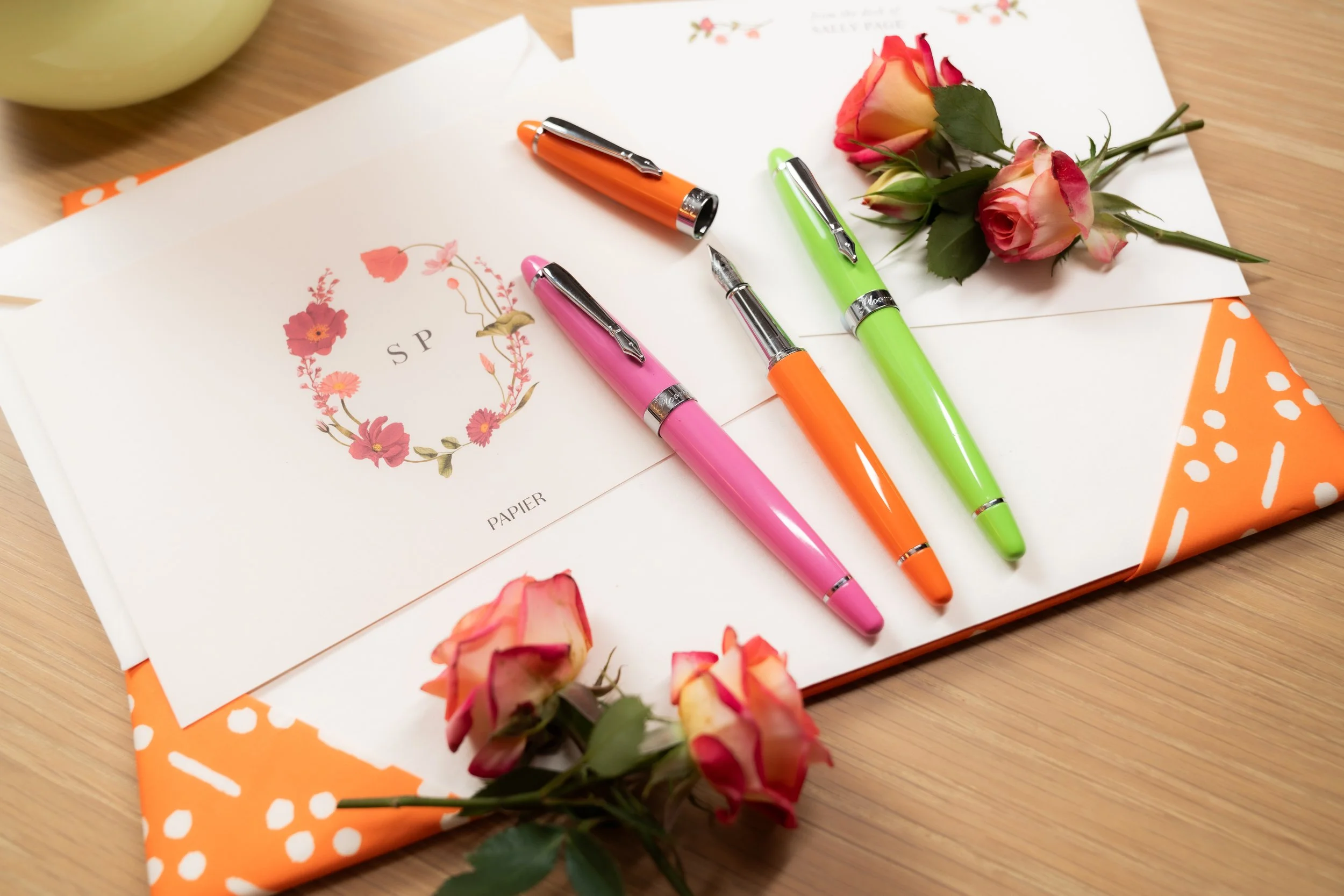 A greeting card with a floral wreath and the letters 'SP' on it, three colorful pens in pink, orange, and green, two pink and cream roses, and a white envelope on an orange polka-dotted wrapping paper on a wooden surface.