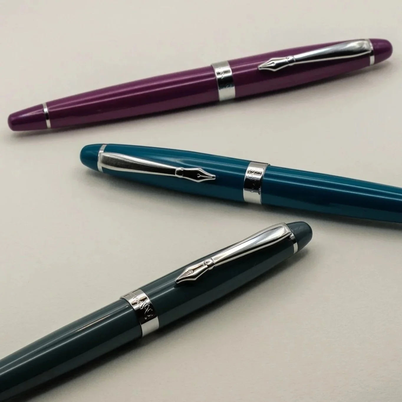 Three pens with silver clip and band, purple, blue, and dark green, arranged on a white surface.