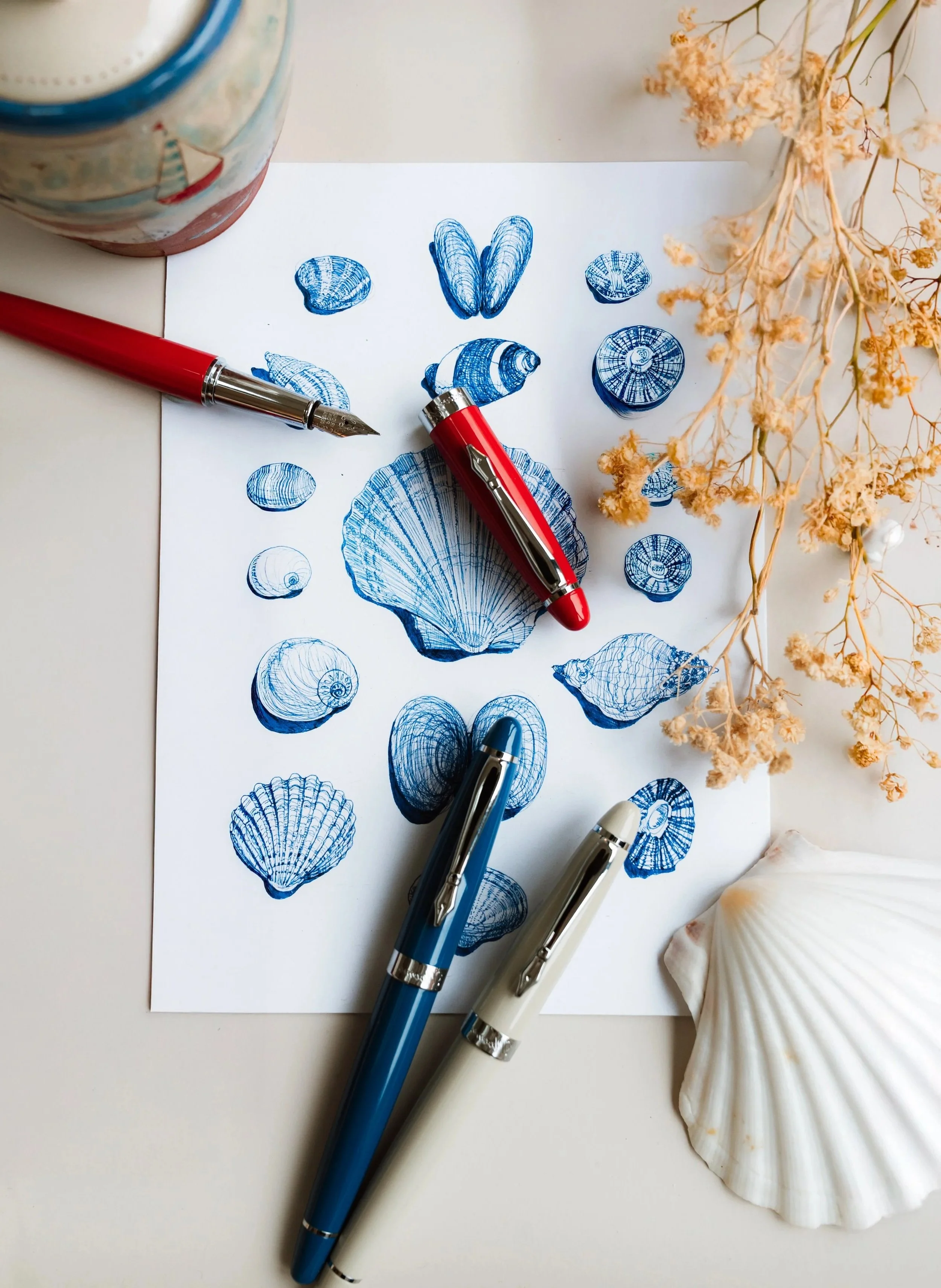 A sheet of paper with blue ink shells drawings, three fountain pens on it, dried flowers, and a shell.