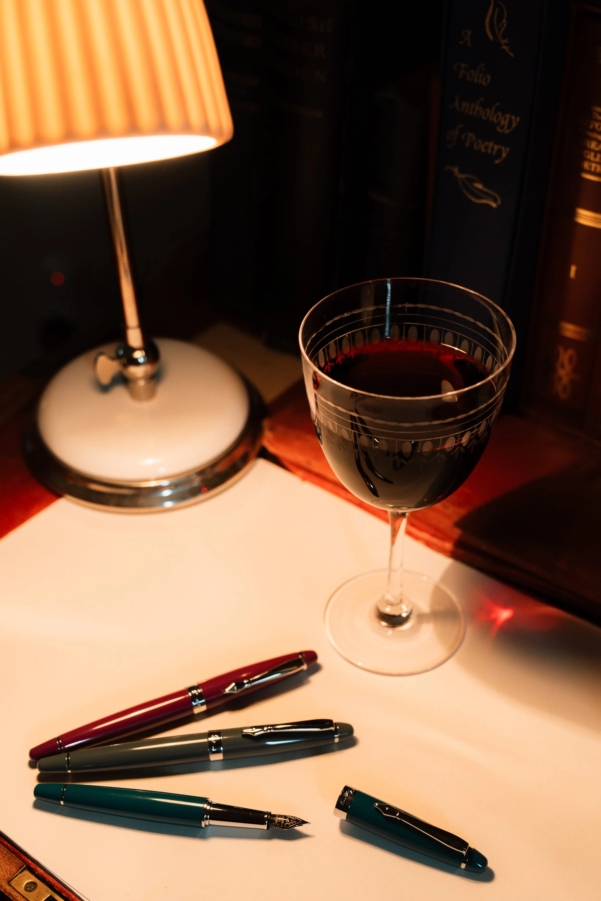 A glass of red wine on a white desk, with three pens and a small lamp, and books on a dark background.