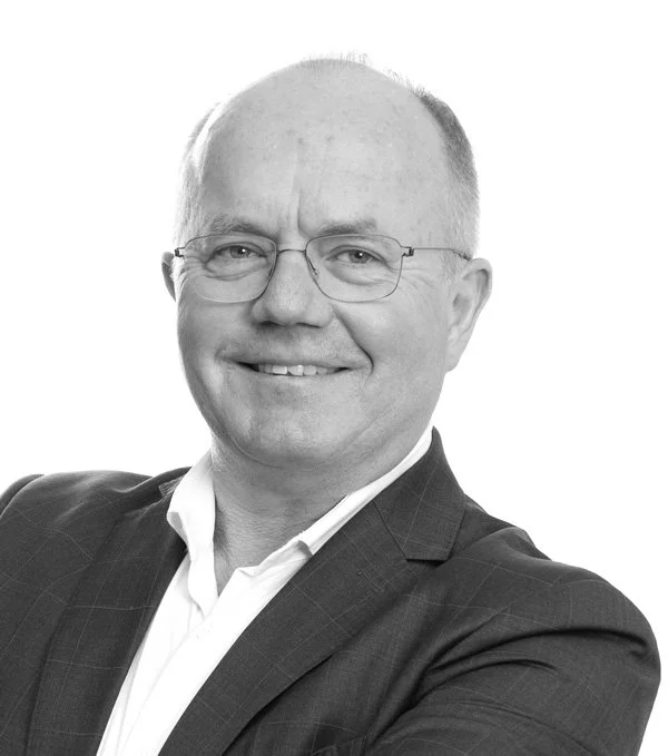Portrett av en mann i dress med briller, smilende, mot en hvit bakgrunn.