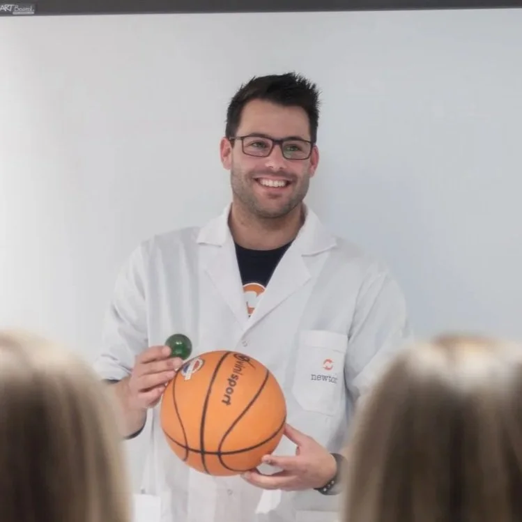 En mann med briller og skjegg smiler, iført hvit lab-frakk, som holder en basketball og en liten grønn ball. Han står foran en hvit tavle og ser ut til å holde en presentasjon.