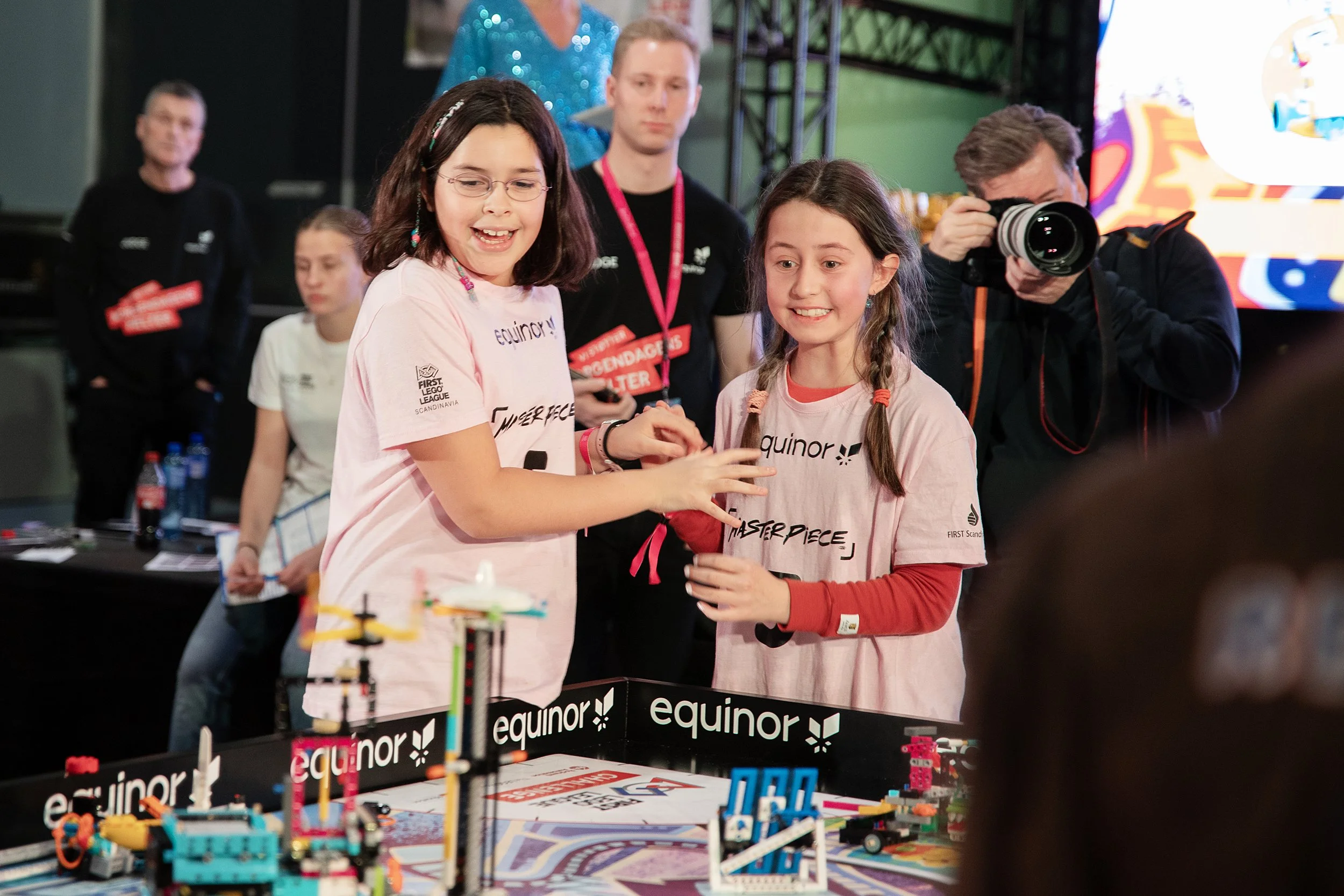 To jenter deltar i en Lego Roboter konkurranse. En voksen fotograferer, mens andre voksne og barn ser på. Konkurransen ser ut til å være i en innendørsarena med branding fra Equinor.