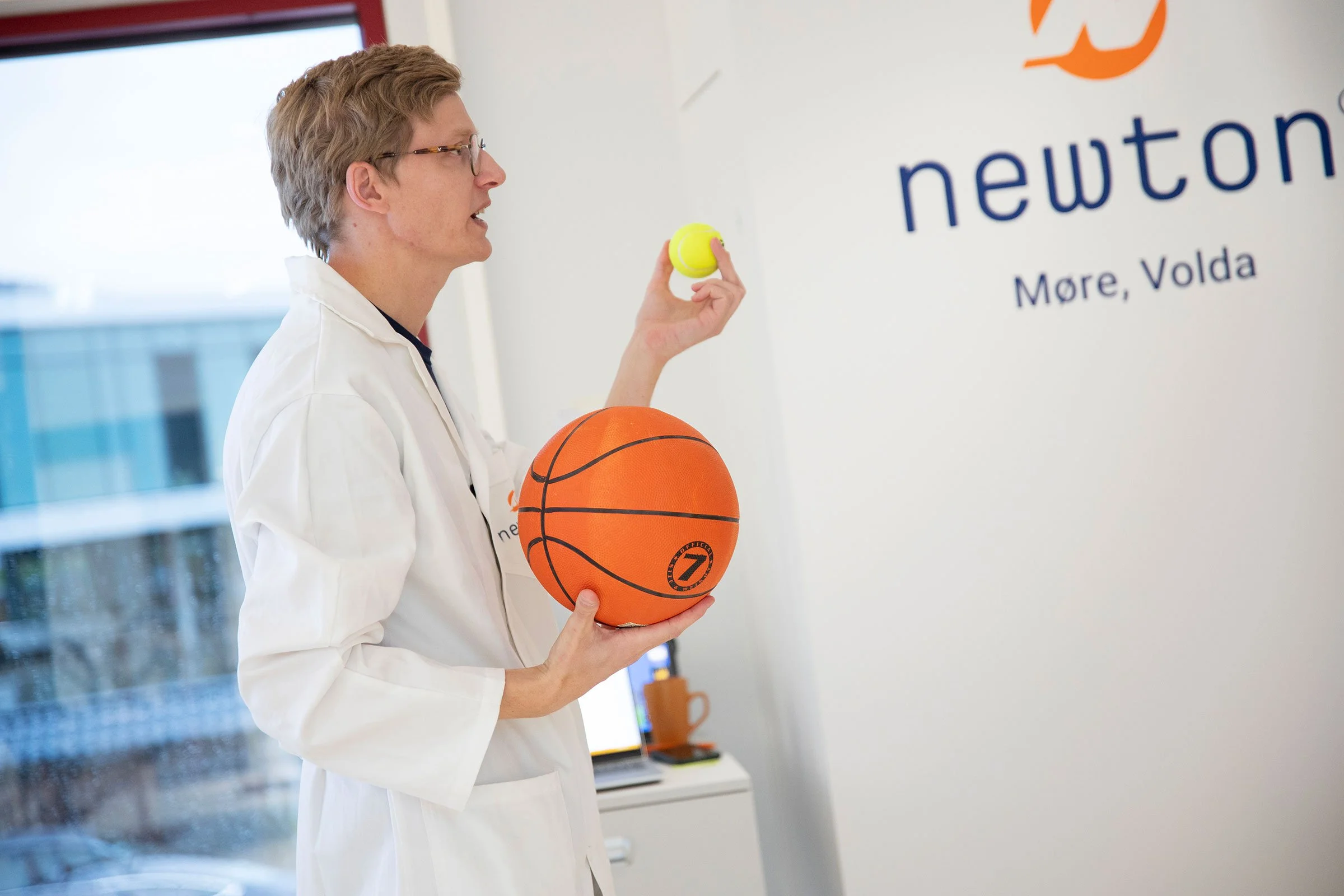 En Newton-lærer i hvit labfrakk holder en basketball og en tennisball, står foran en vegg med logoen til Newton Møre, Volda.