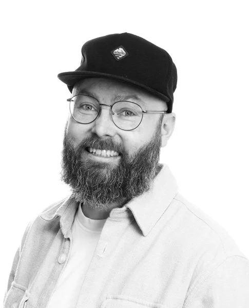 Portrett av en mann med skjegg, briller og svart caps, smilende, i svart-hvitt.