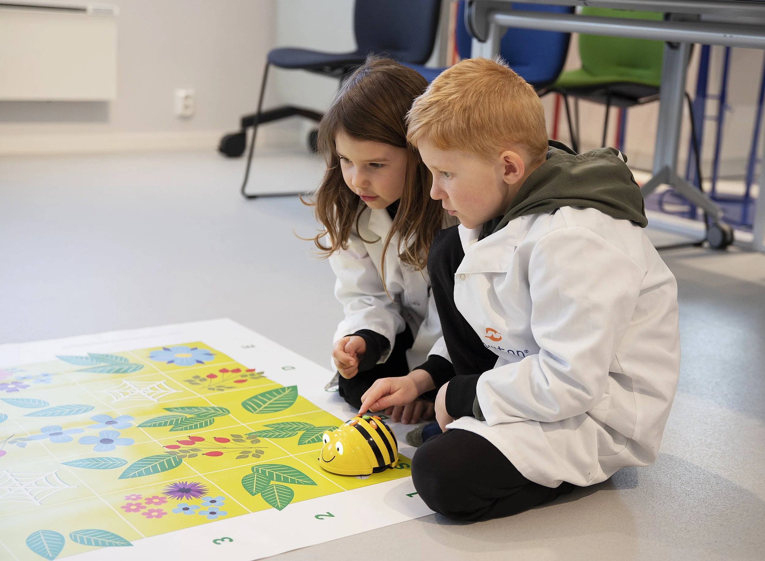 To barn, en jente med brunt hår og en gutt med blond hår, leker med en robot på et bord med fargeglade plakater med blomster og blad på. De er kledd i hvite smocker. Bakgrunnen viser stoler og et bord.