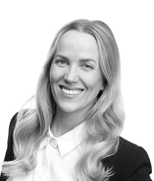 Profesjonell portrett av en smilende kvinne med langt, blond hår iført sort jakke og hvit skjorte, fotografi i svart-hvitt.