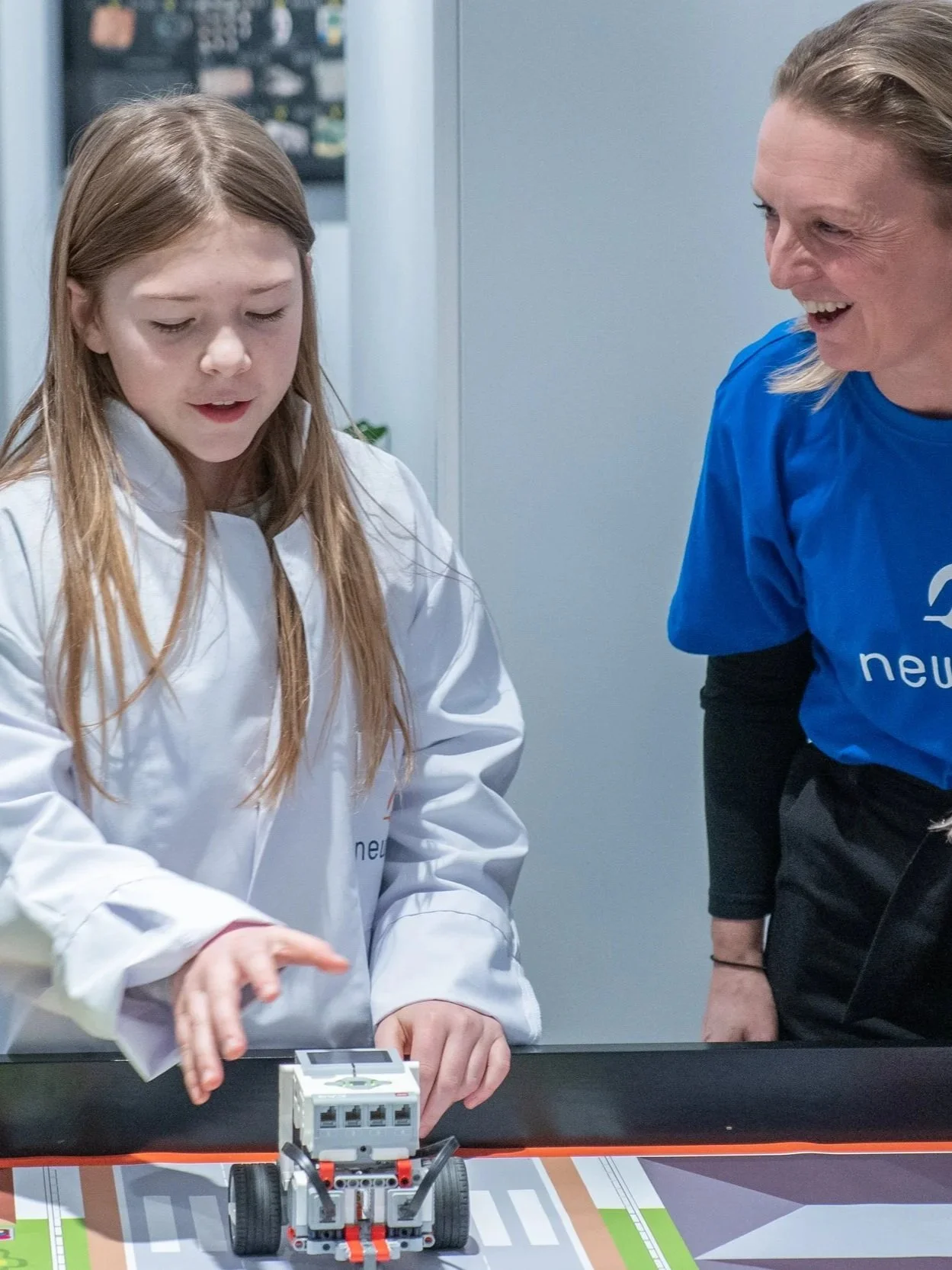 En jente i hvit labfrakk og en voksen i blå t-skjorte ser på en LEGO robot som kjører på et plan med ulike fargede områder.