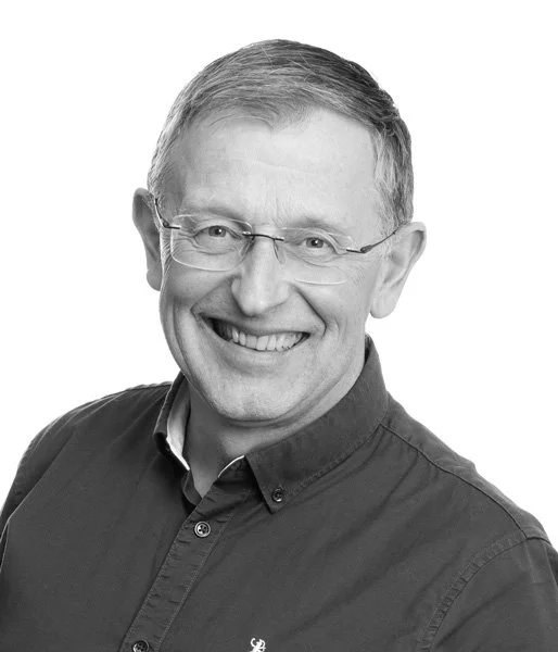 En smilende mann med brunt hår, briller og en sort skjorte, fotografert i svart-hvitt.
