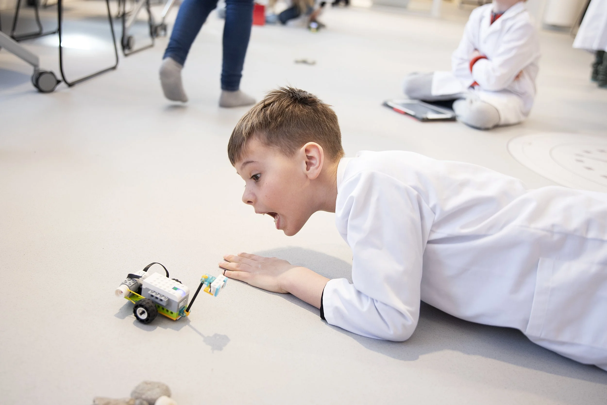 En gutt i hvit labfrakk leker med en liten robot på gulvet. Det er andre personer i bakgrunnen, noen av dem står og sitter.
