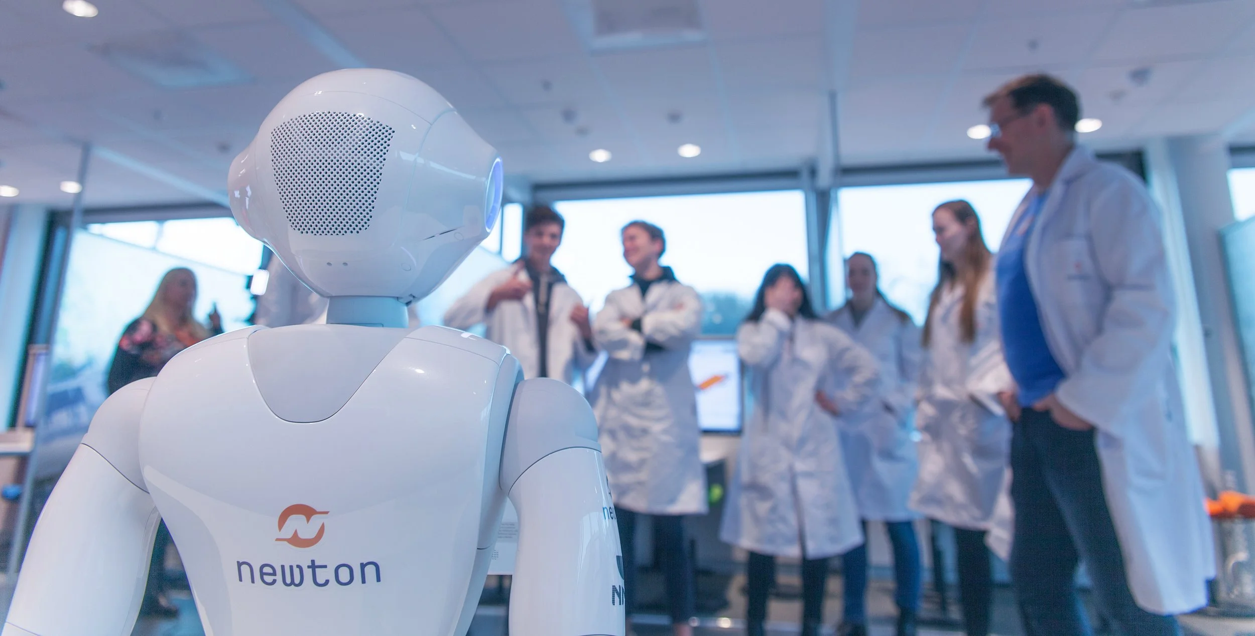 En robot med merket 'Newton' står foran en gruppe mennesker i laboratoriefrakker som smiler og prater.