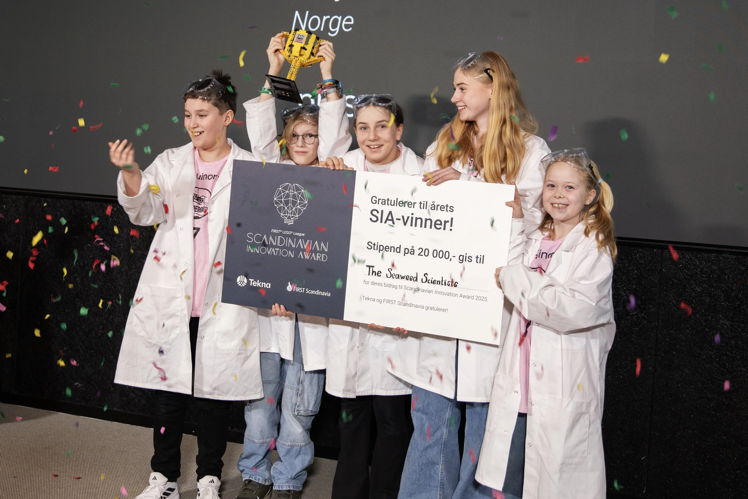 Gruppe barn i hvite labfrakker feirer med konfetti, hvor en av dem holder opp en LEGO trofé og et stort sjekk som gratulerer vinnerne av Scandinavian Innovation Award 2025.