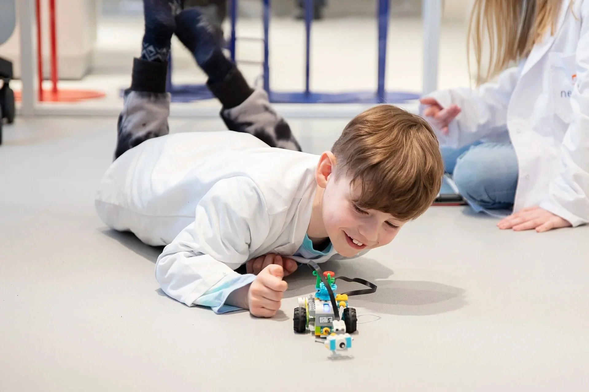 Ung gutt i laborklær som legger seg på gulvet, spiller med en liten robot laget av byggeklosser. En person i hvit frakk er i bakgrunnen.